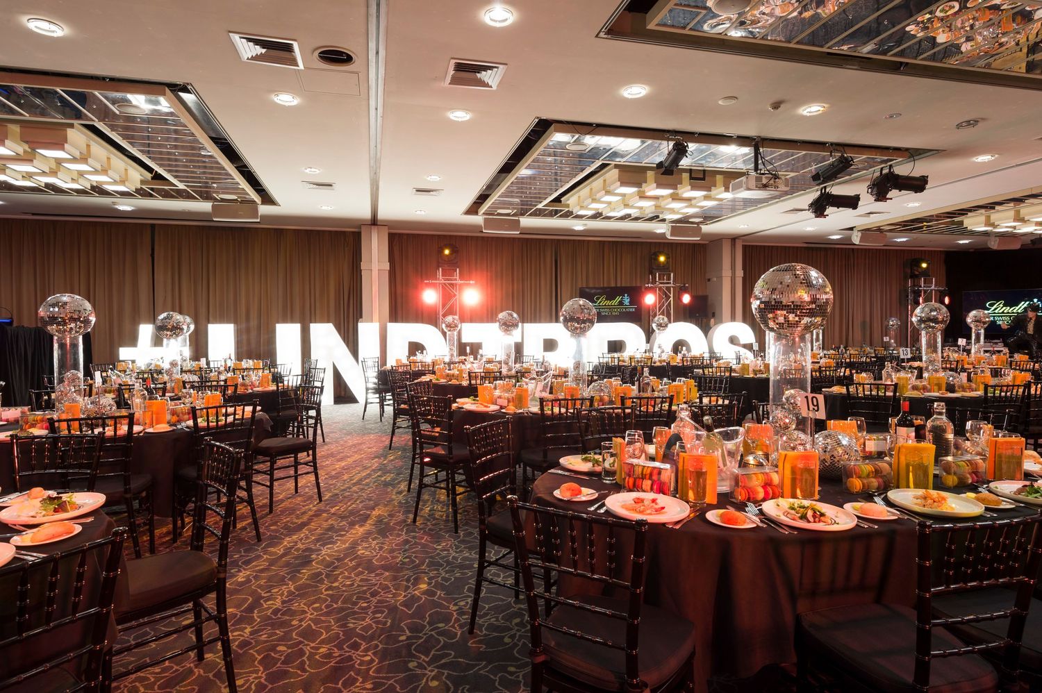 Elegant banquet hall setup with round tables decorated in orange linens and centerpieces under ornate ceiling lighting.