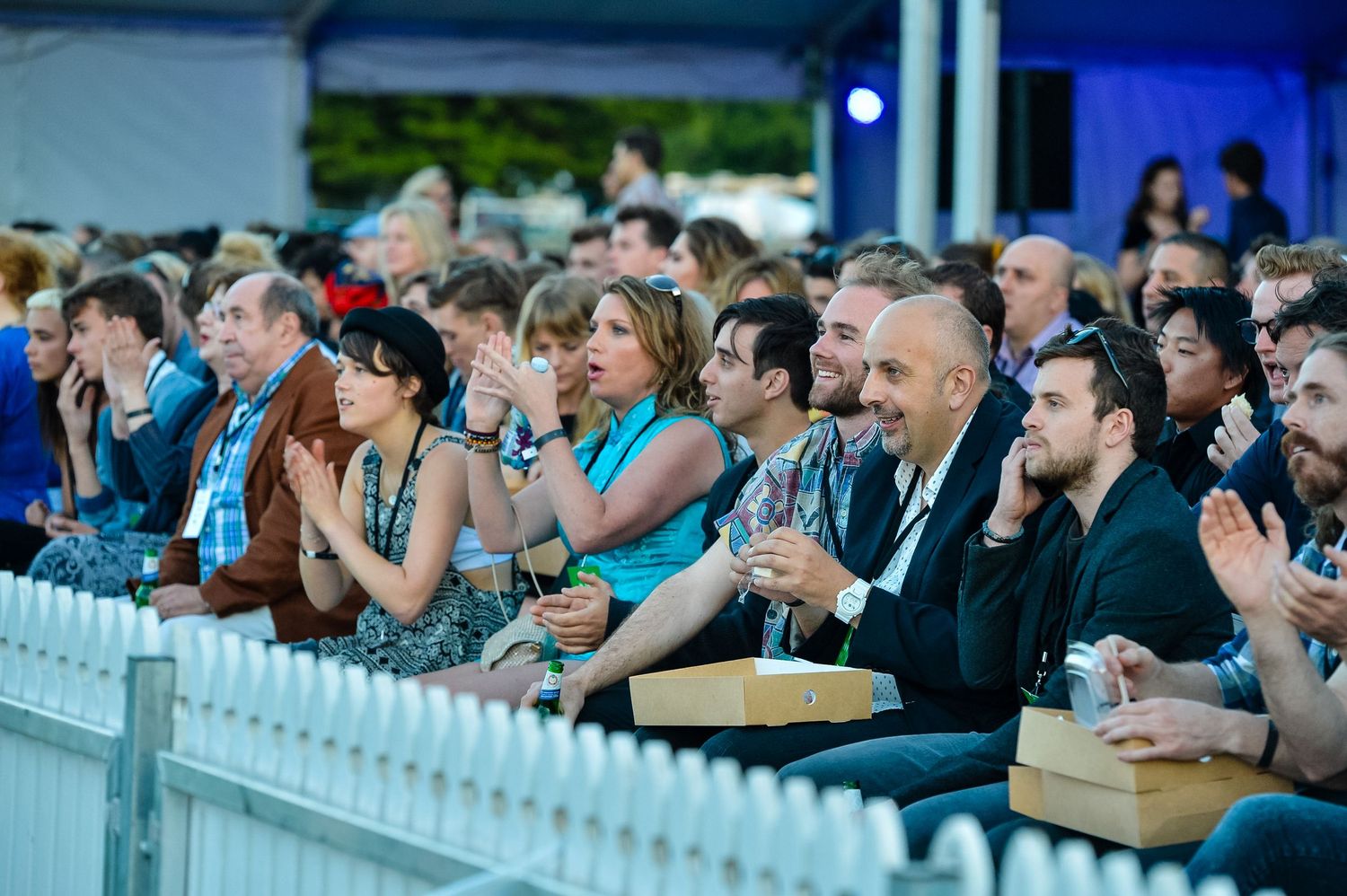 Wedding guests watch ceremony from white picket fence seating area at outdoor venue.