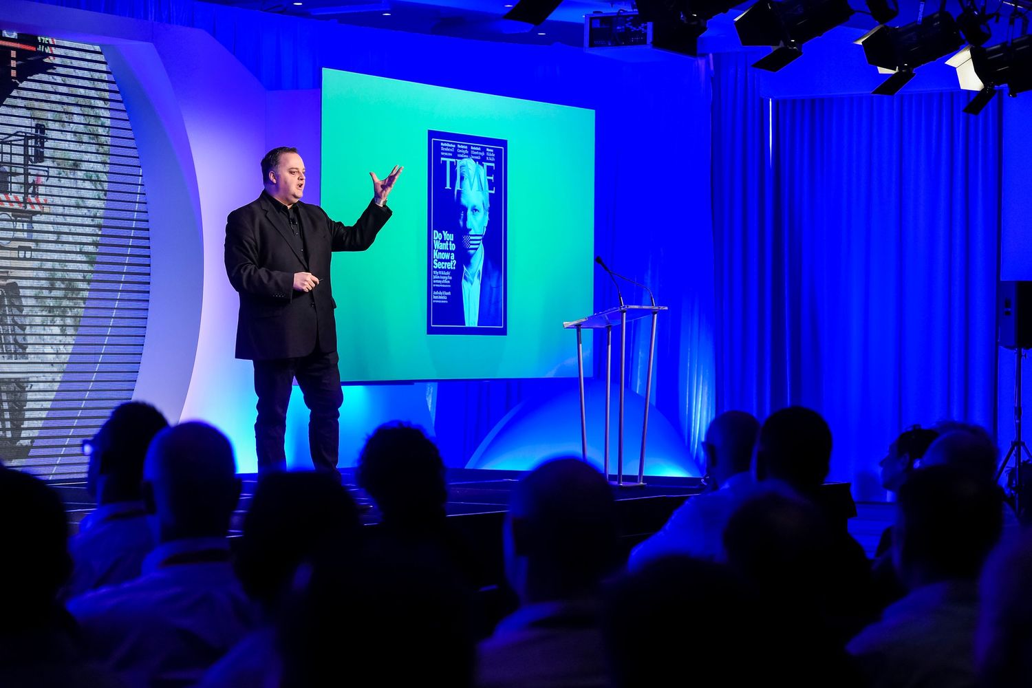 Large conference hall with dramatic blue stage lighting and projection screens during presentation.
