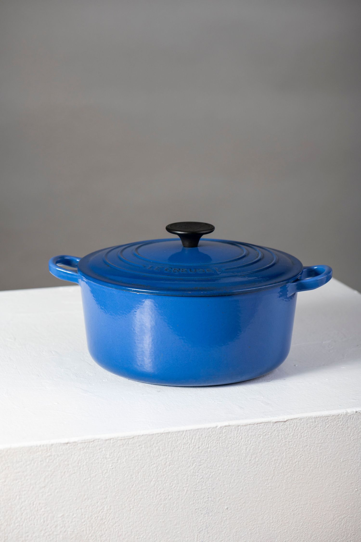 Four identical blue enameled cast iron braising pans with black lids arranged in a row on a white surface.
