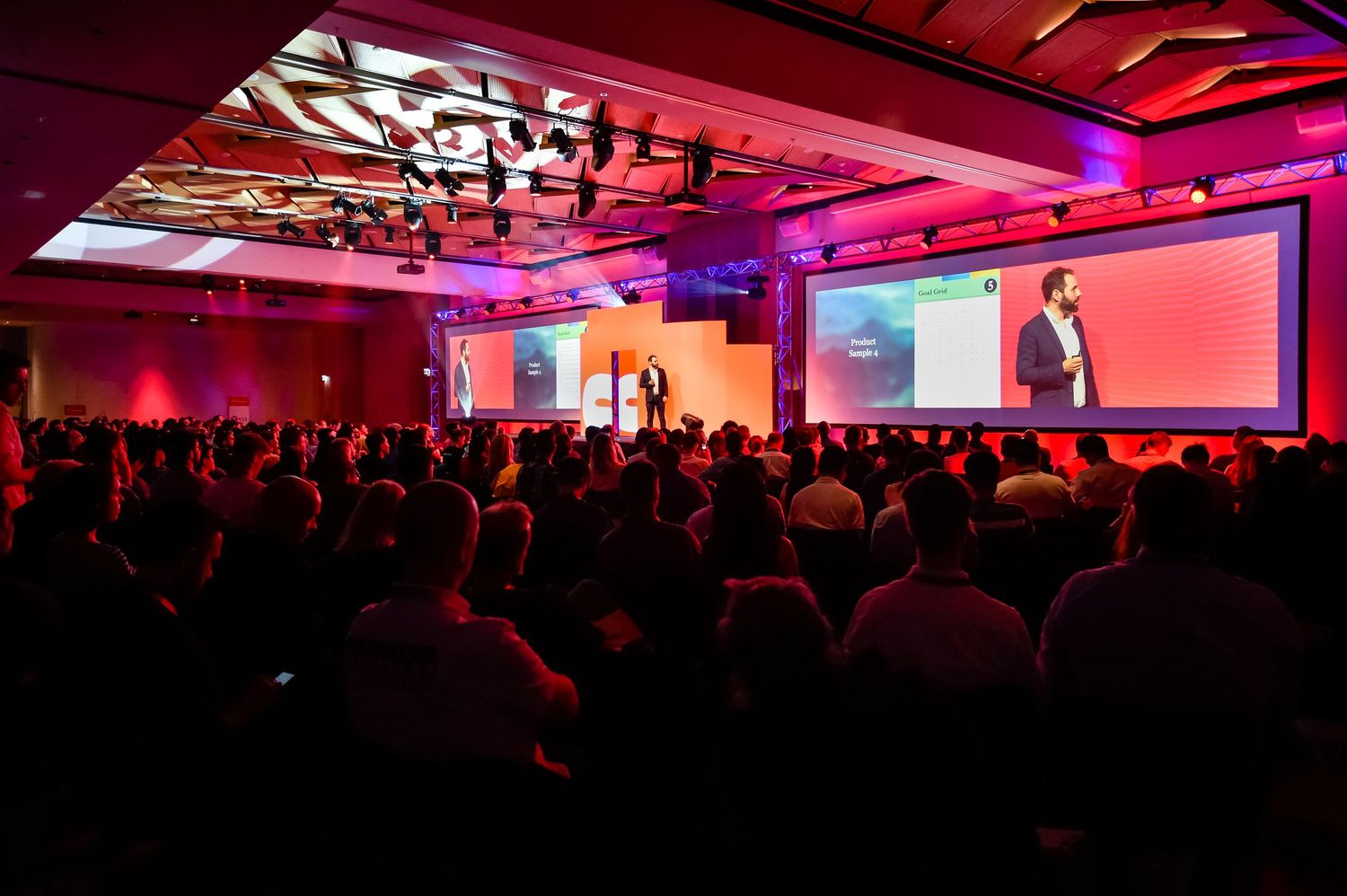 Large audience watches presentation in dimly lit conference hall with red ambient lighting.