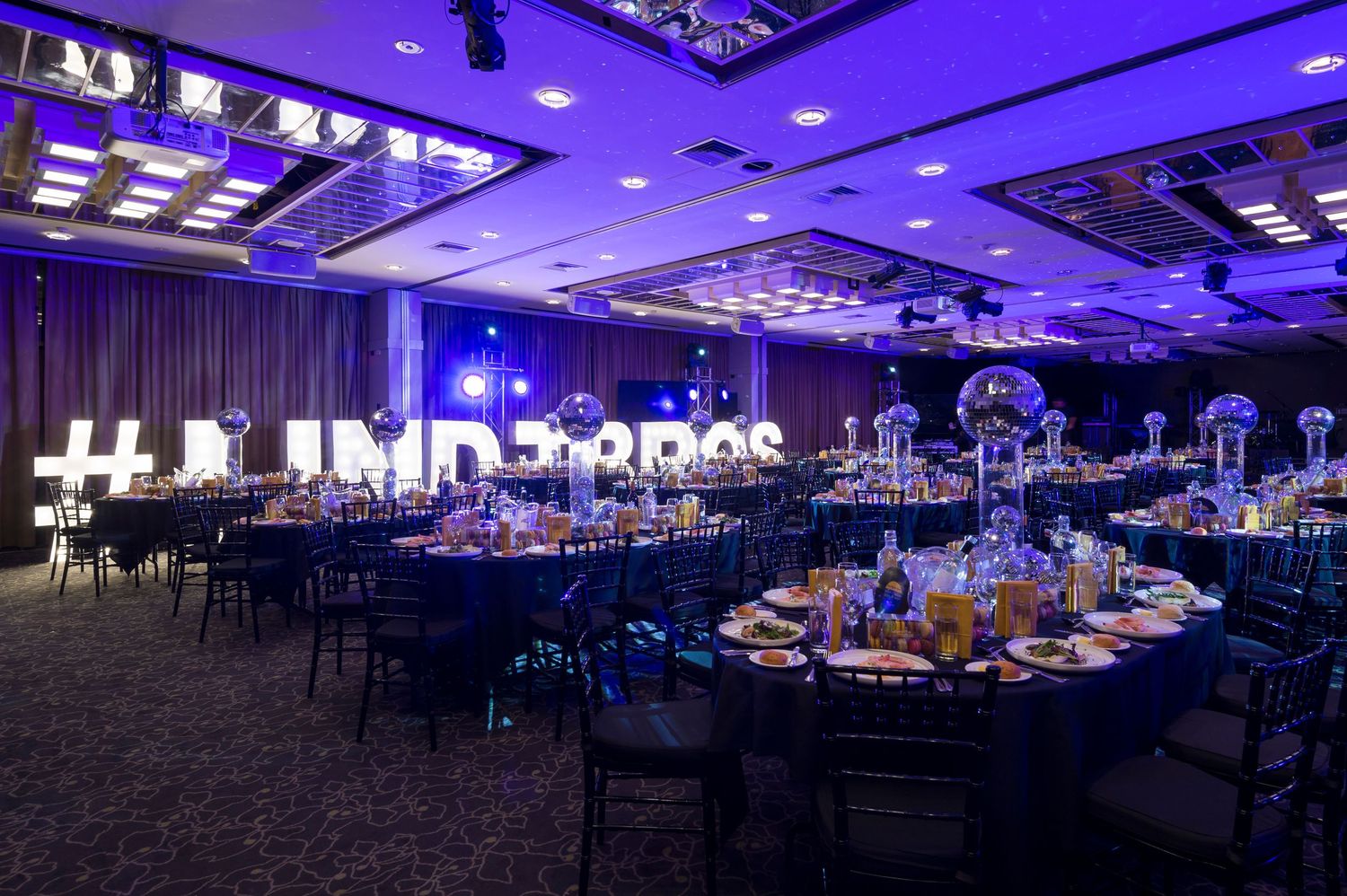 Large corporate event space with purple ambient lighting illuminating multiple round tables and chairs in a ballroom setting.