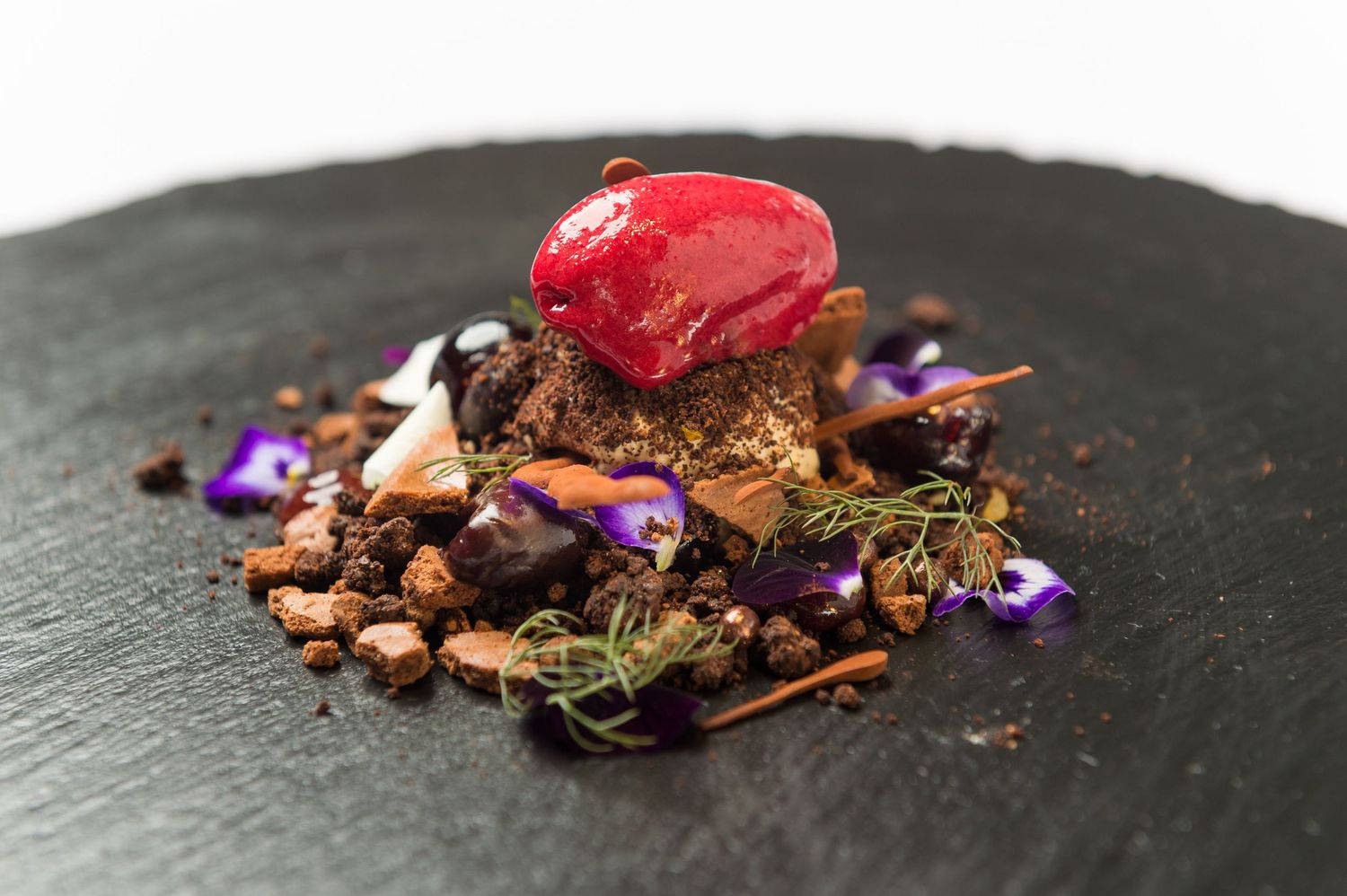 Artistic plating of chocolate and red elements on a black slate surface with edible flowers.
