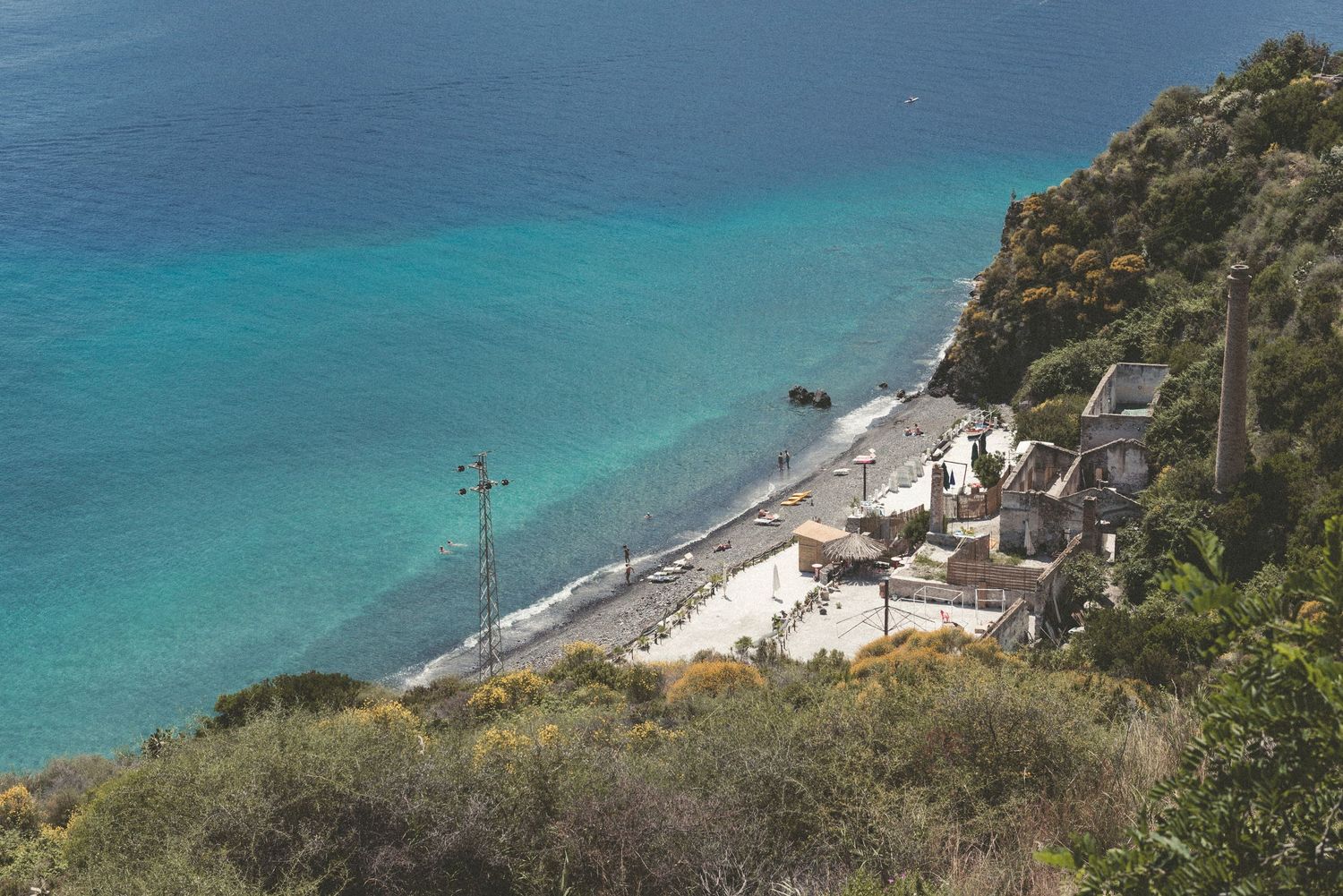 Aerial panoramic view of turquoise Mediterranean coastline with rocky cliffs and winding coastal road.
