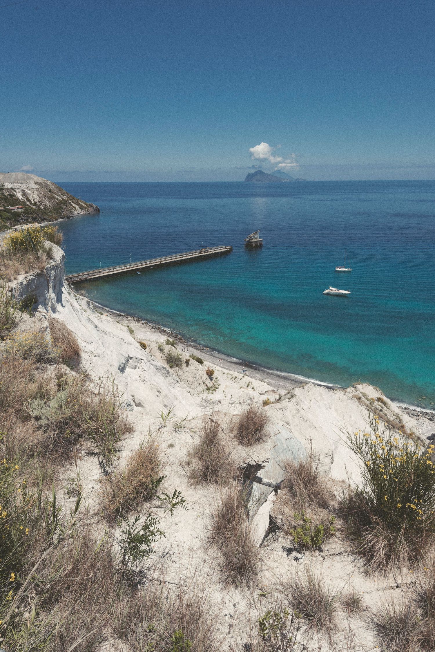 Scenic Mediterranean coastline with white cliffs, turquoise waters, and a long pier extending into the sea near distant islands.