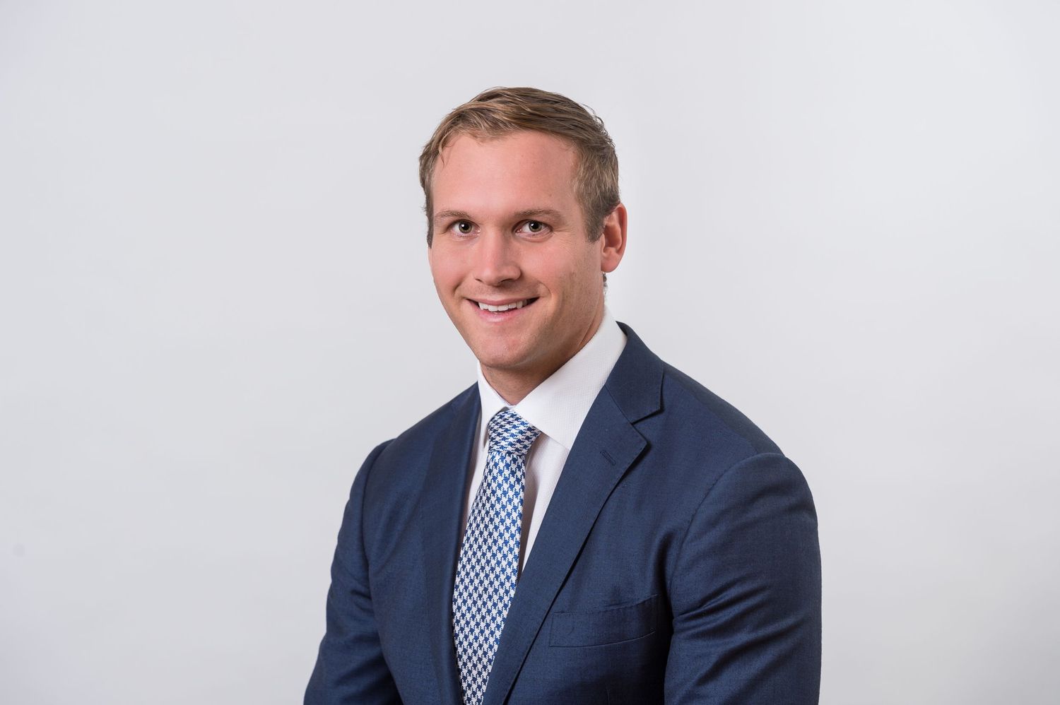 Professional in navy suit and blue tie smiles against white background.