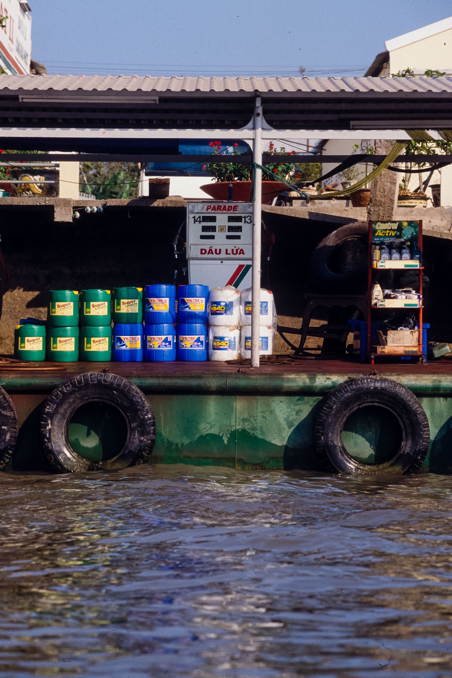 Colorful oil drums and tires line a waterfront storage area.