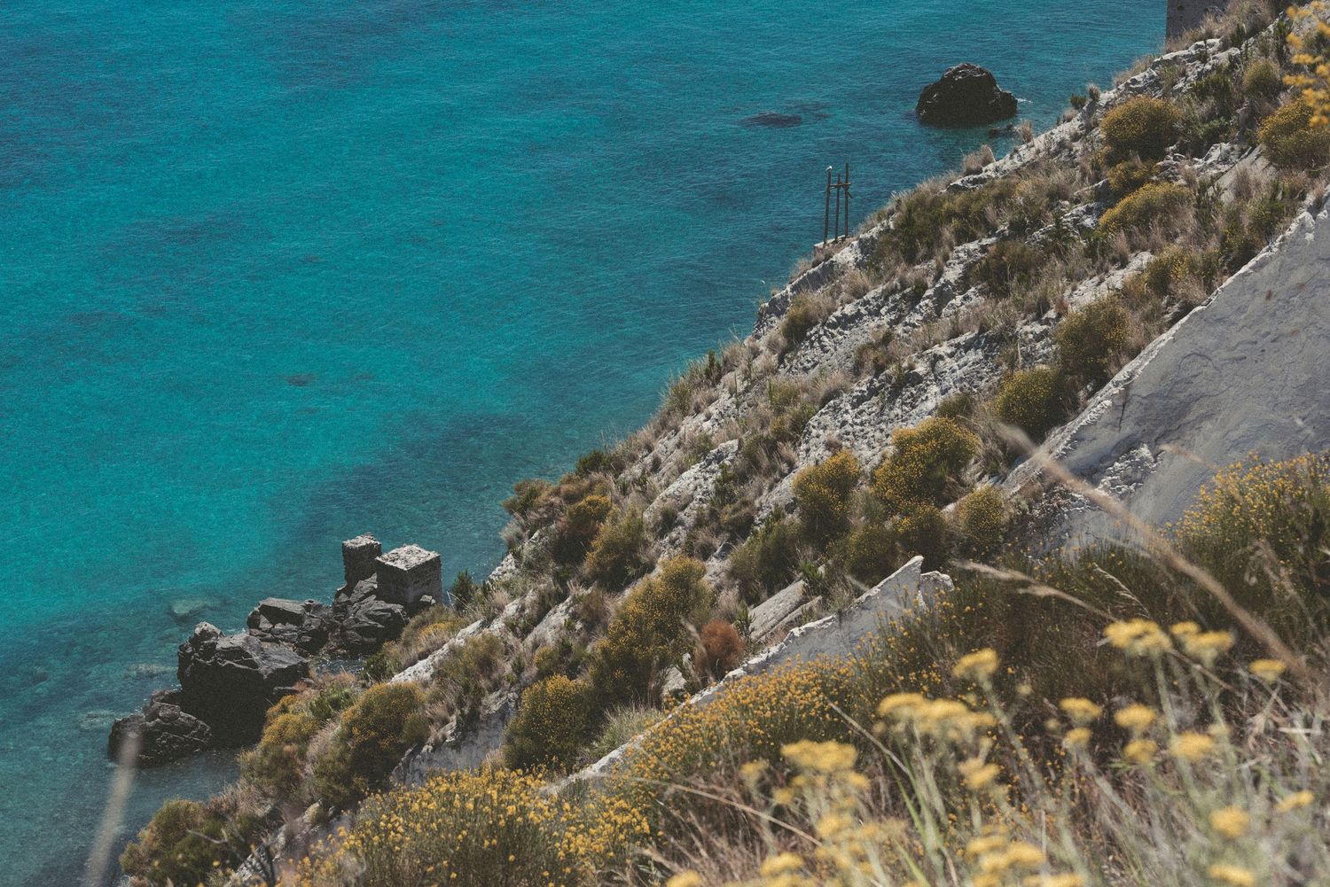 Coastal cliff overlooking turquoise Mediterranean waters with rugged terrain and winding road path.