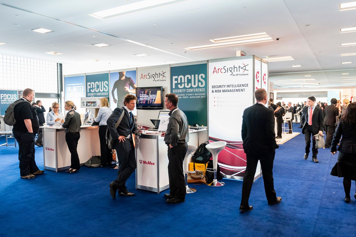 Exhibition hall with white booths and blue carpeting showing attendees networking at a trade show.