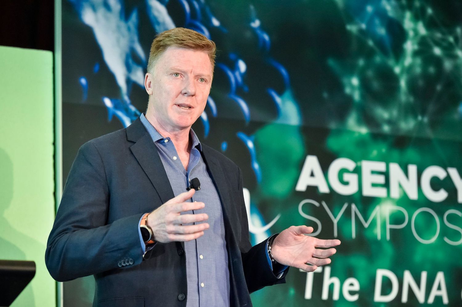 Speaker presenting on stage with DNA-themed digital backdrop at a business symposium.