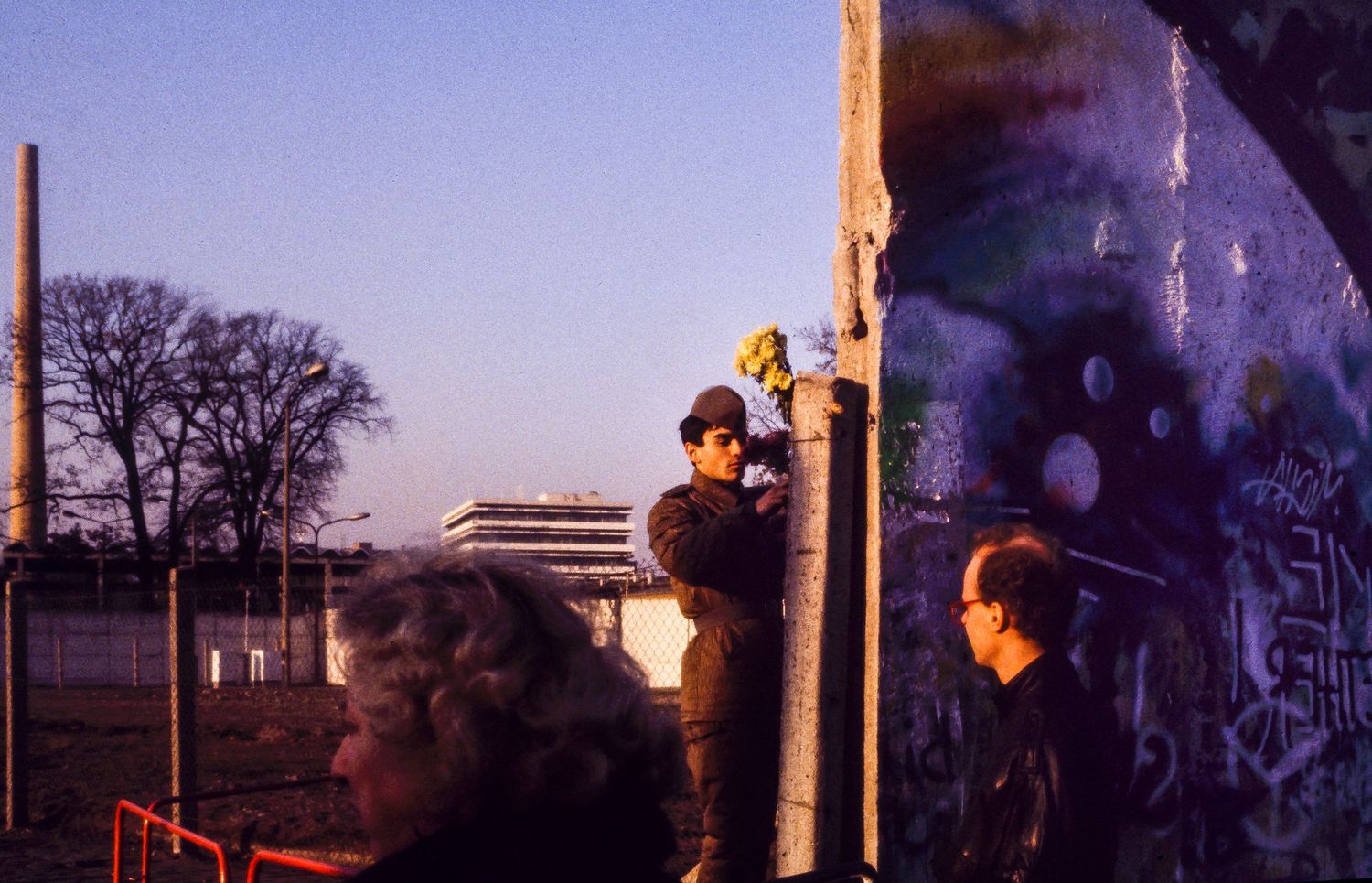 Silhouettes of people walking along the Berlin Wall at sunset with graffiti visible on the concrete barrier.