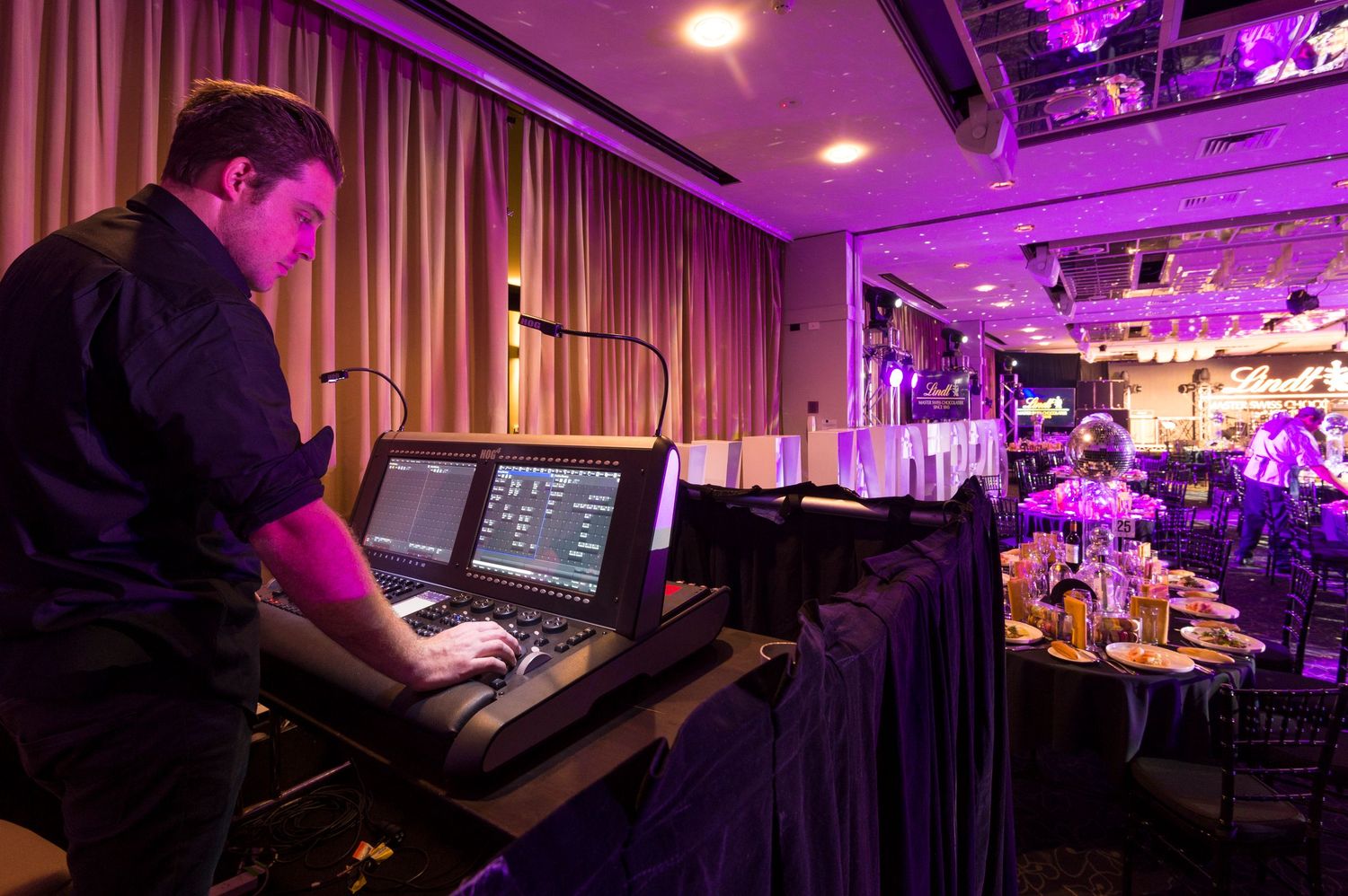 Sound technician operates digital mixing console at venue with purple stage lighting and curtained backdrop.