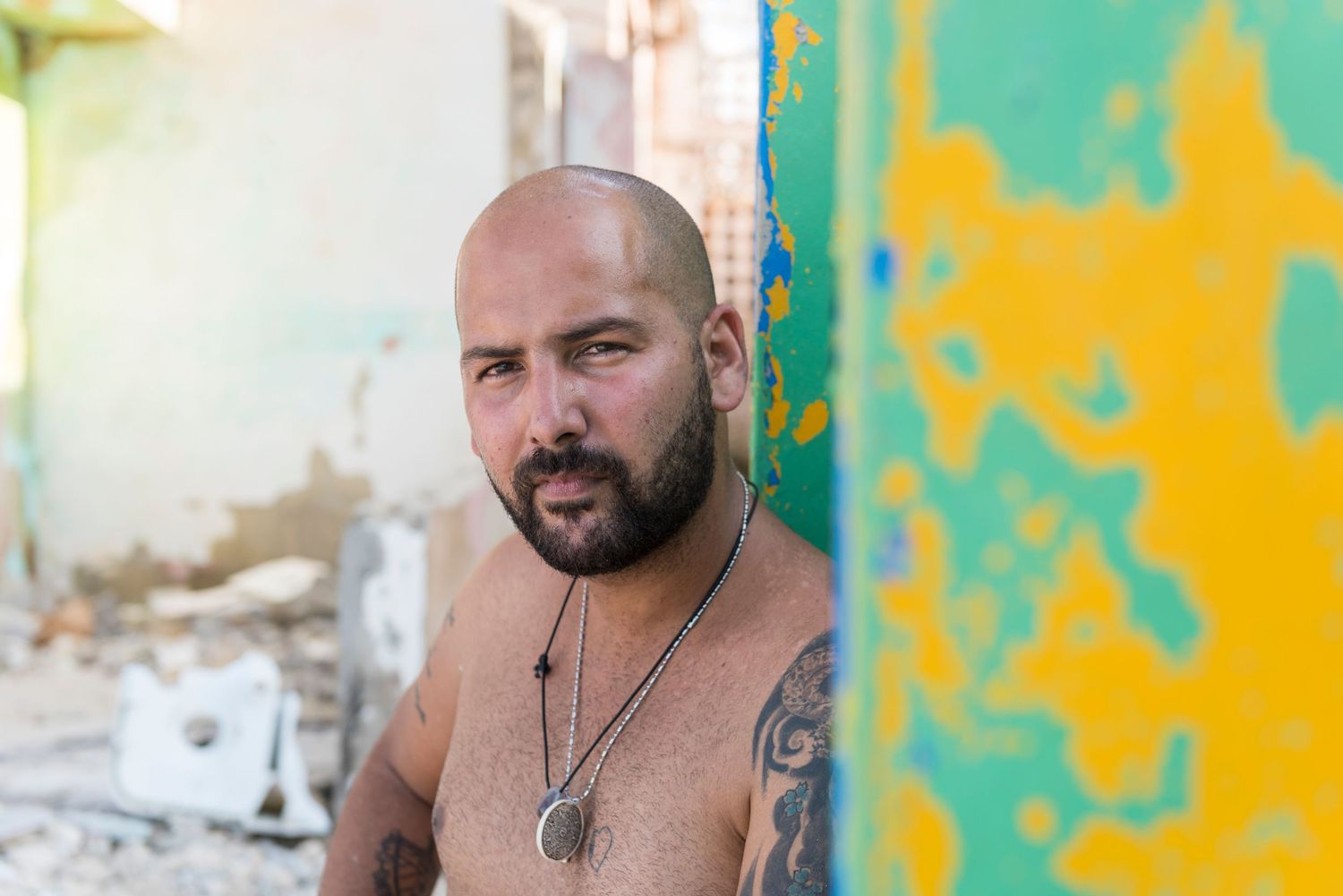 Shirtless man with tattoos and necklace poses against colorful wall with graffiti.