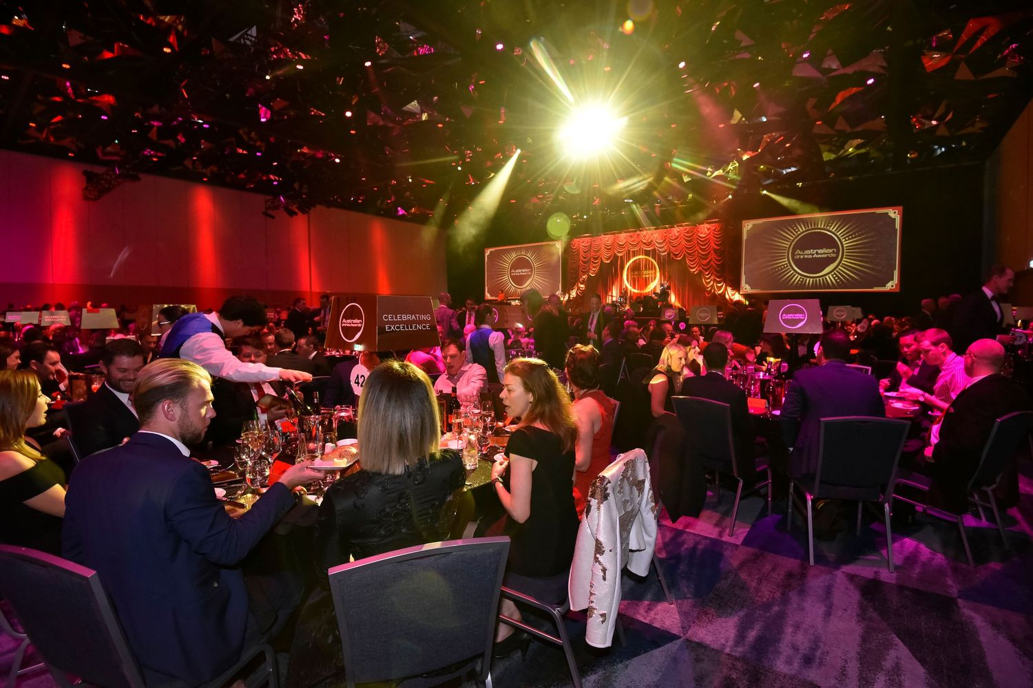 Guests enjoy an elegant evening gala event with dramatic red lighting and a bright spotlight in an upscale venue.