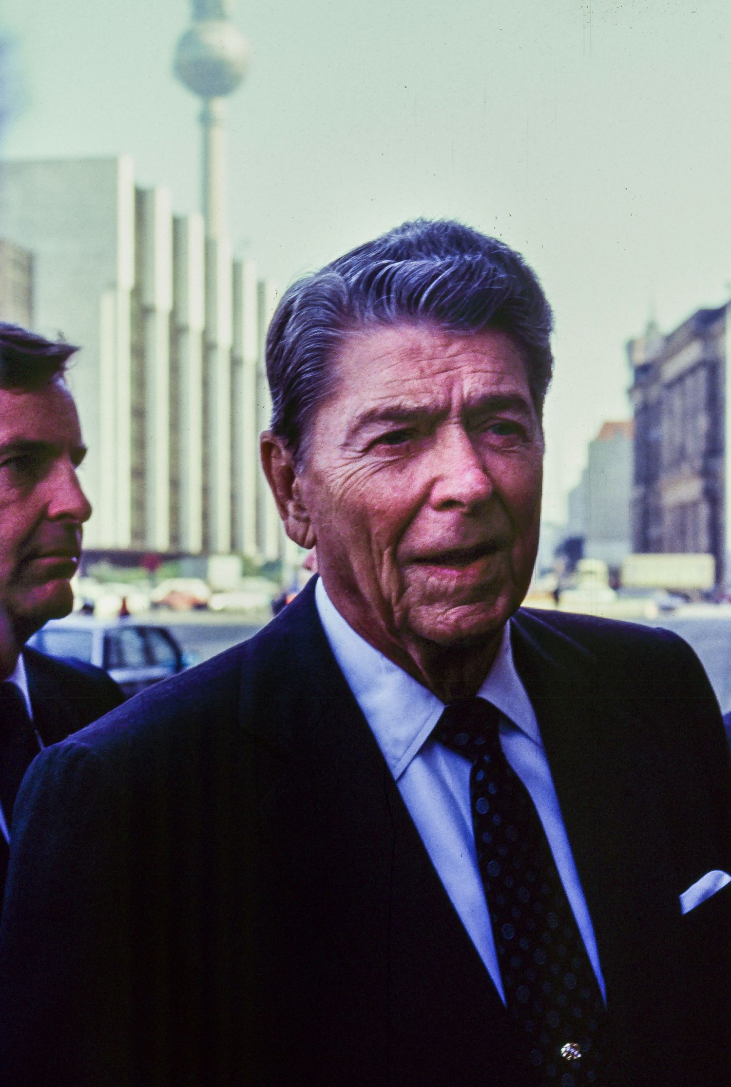 A man in a dark suit photographed in Berlin with the TV Tower visible in the background.