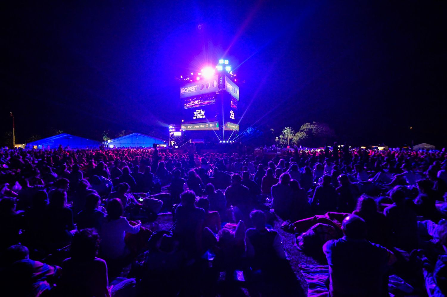 A large crowd gathers at night beneath a glowing blue and purple DJ booth tower at an outdoor music festival.