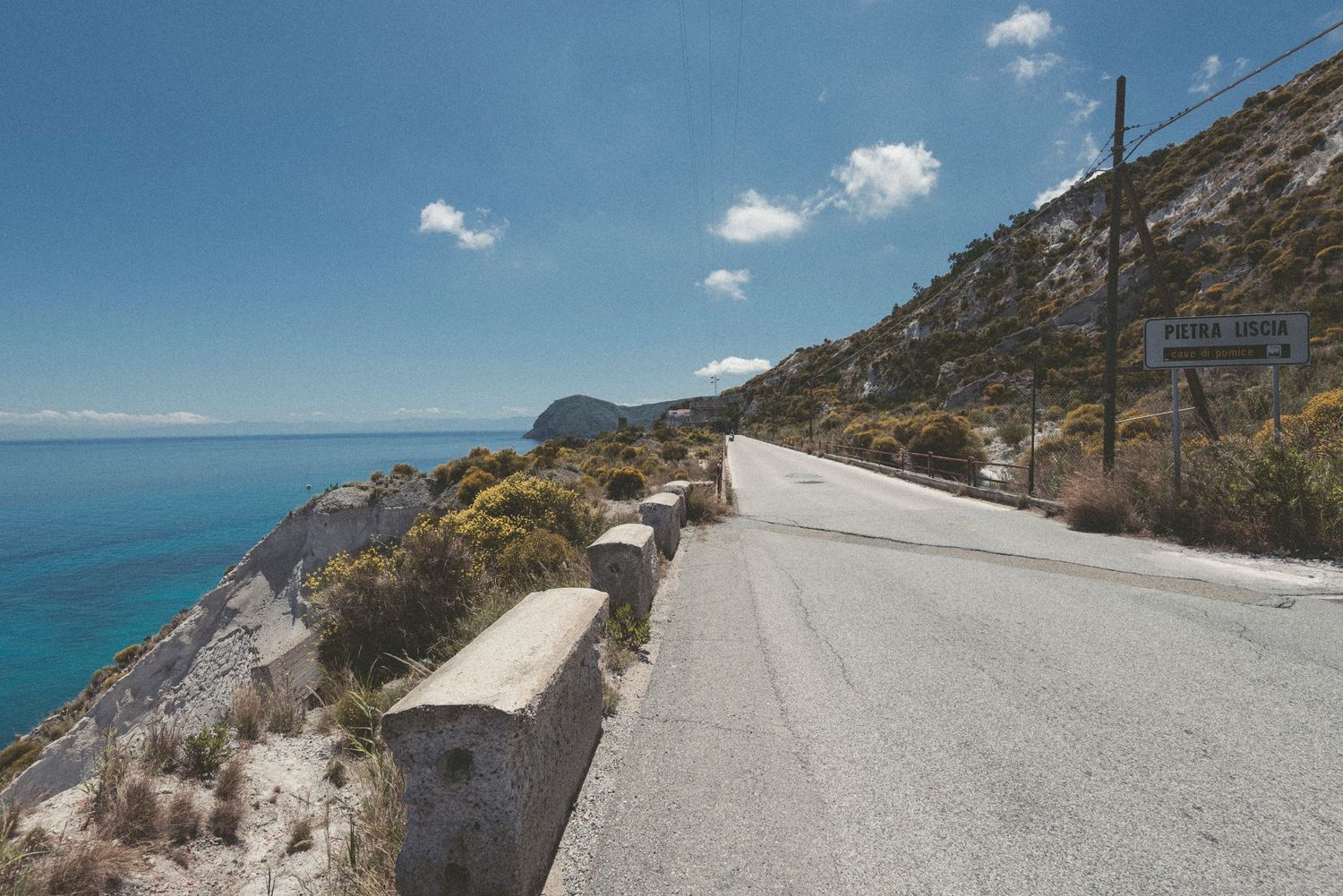 Winding coastal road curves along steep cliffs overlooking the Mediterranean Sea with blue skies and scattered clouds.