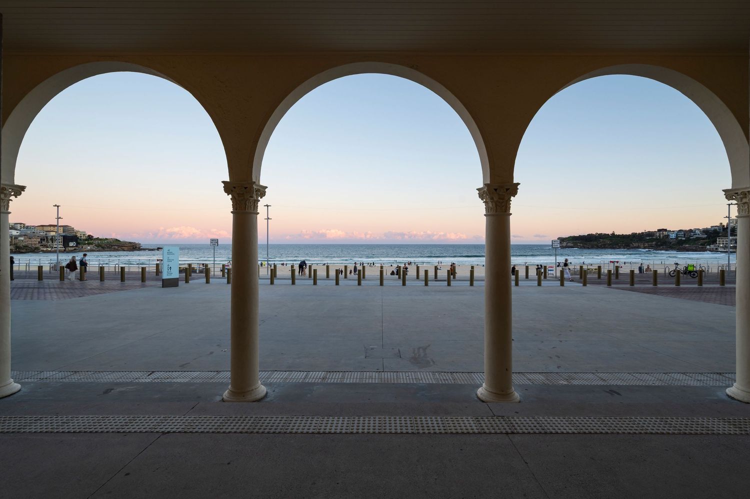 Three arches frame a scenic view of Bondi Beach boardwalk at sunset with the panoramic views ocean and beach in distance.