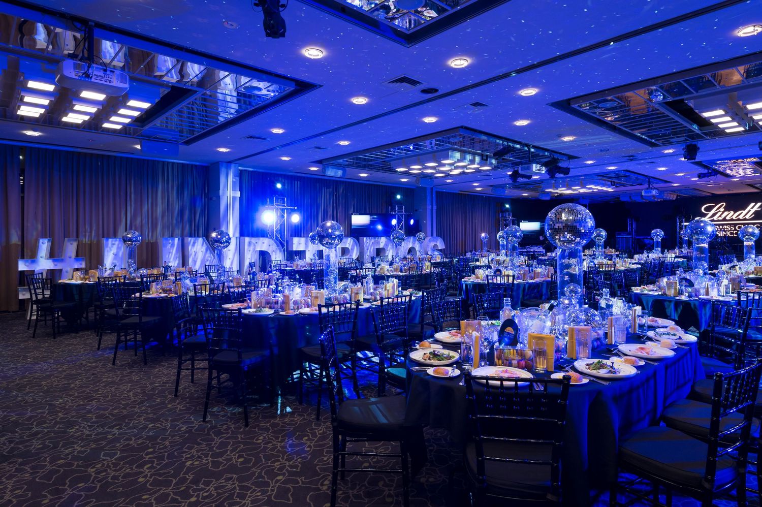 Large event space illuminated in vibrant blue lighting with elegant table settings and modern decor throughout the venue.