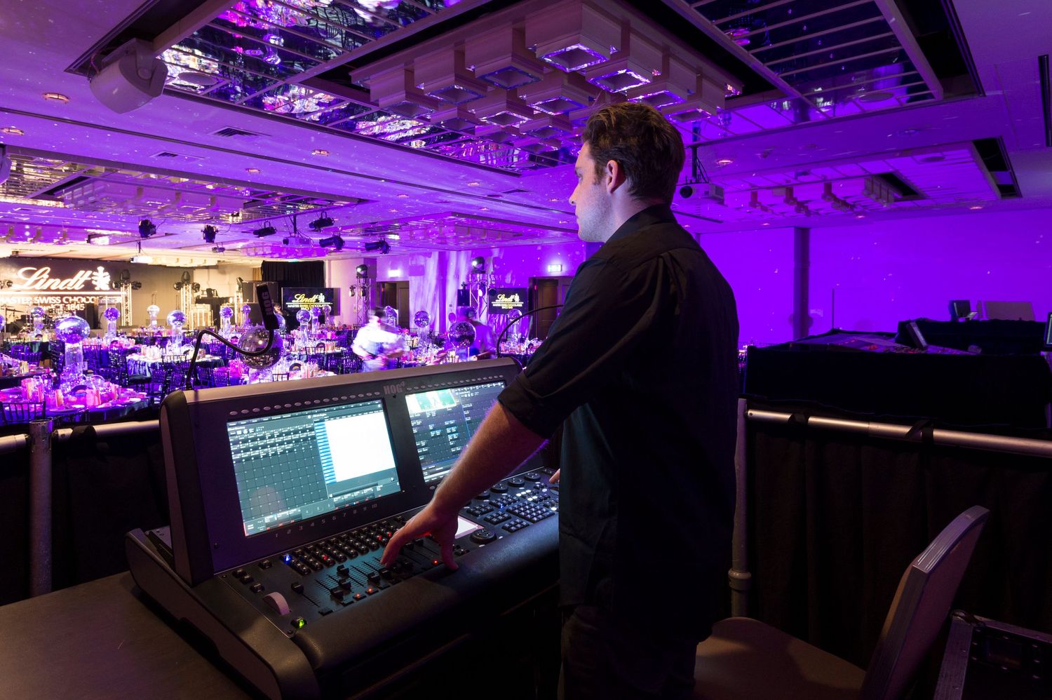 A purple-lit conference or event space with technical equipment and silhouetted figures at control stations.