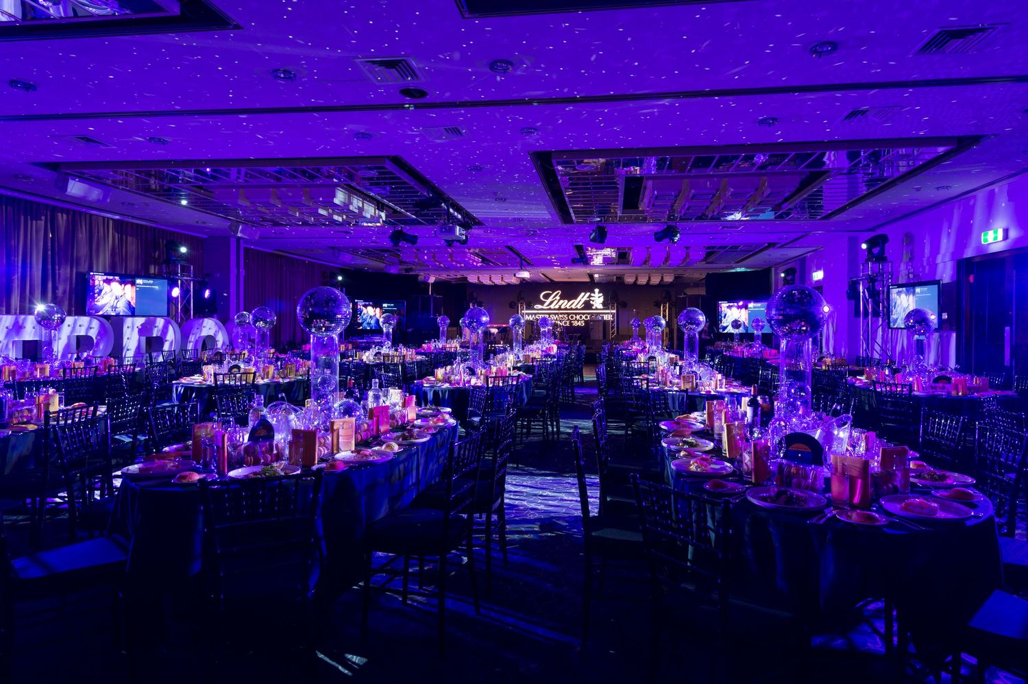 Large event space illuminated in vibrant purple lighting with tables and chairs arranged for a formal gathering.