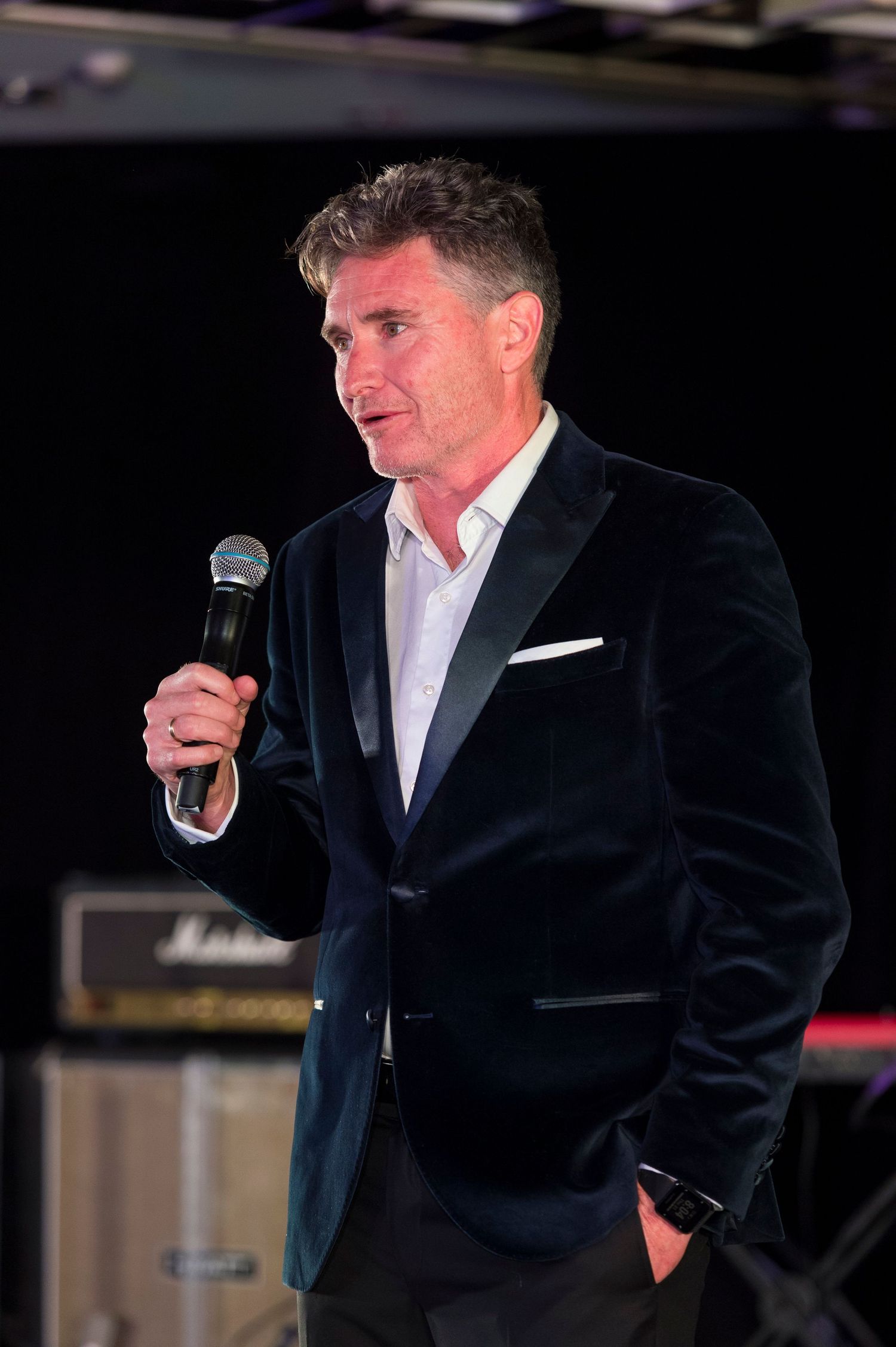 A speaker in a dark velvet blazer and white shirt holds a microphone while presenting at an event.