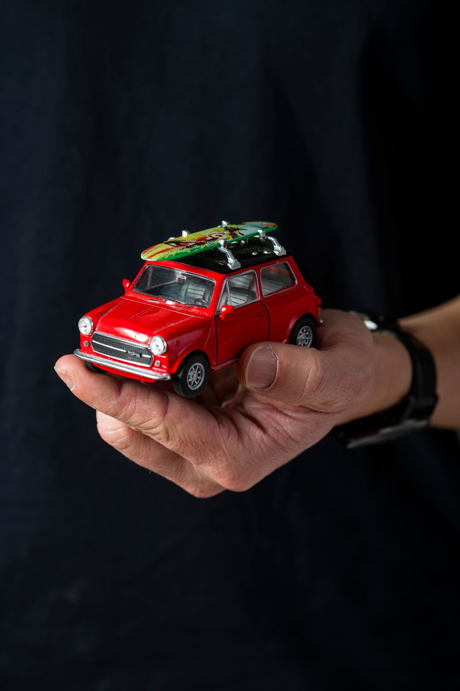 Three hands holding red and green vintage Mini Cooper toy cars against a black background.