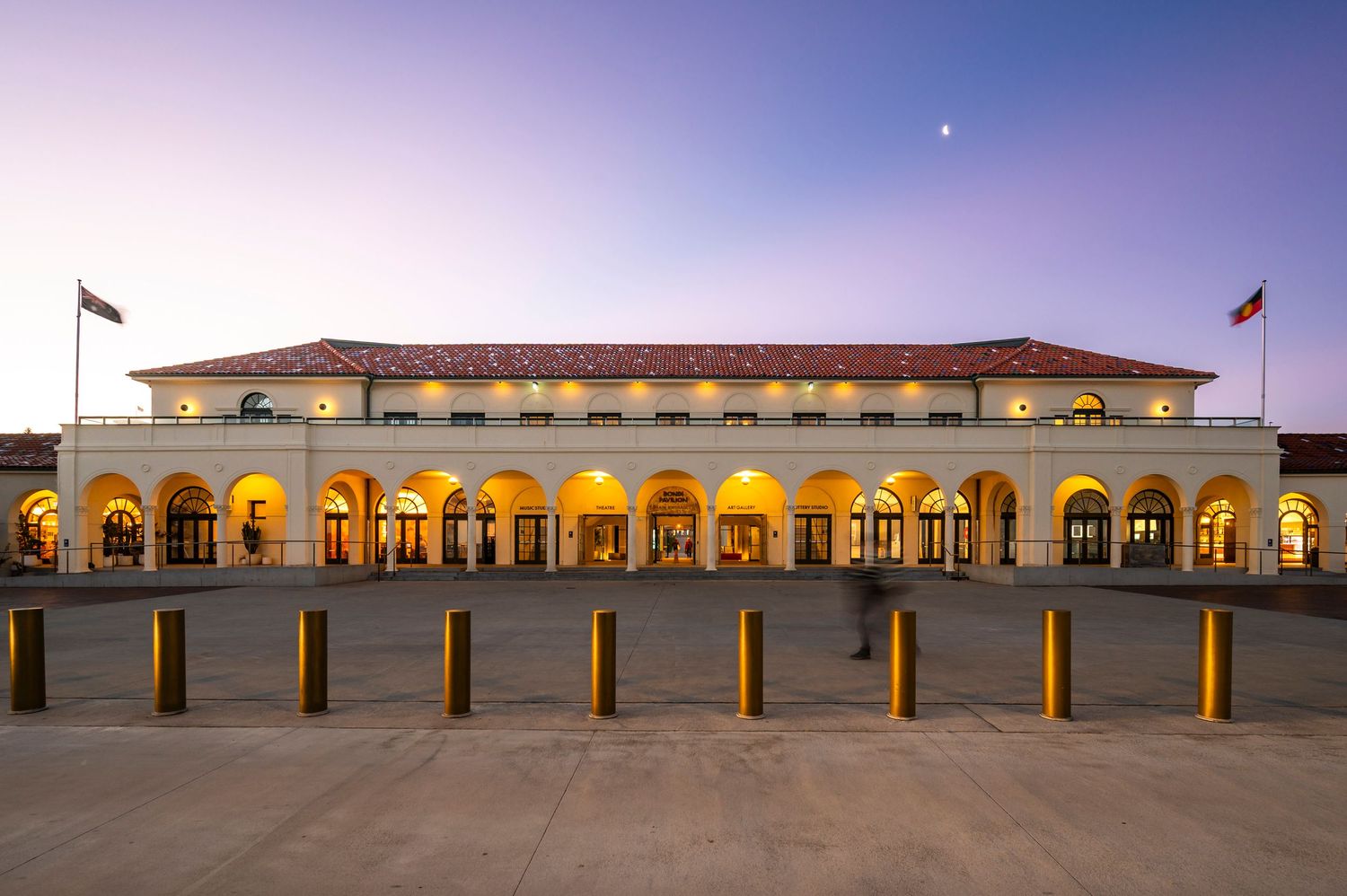 Bondi Pavilion building beach facing with illuminated arches and bollards glows warmly at dusk under a purple sky.