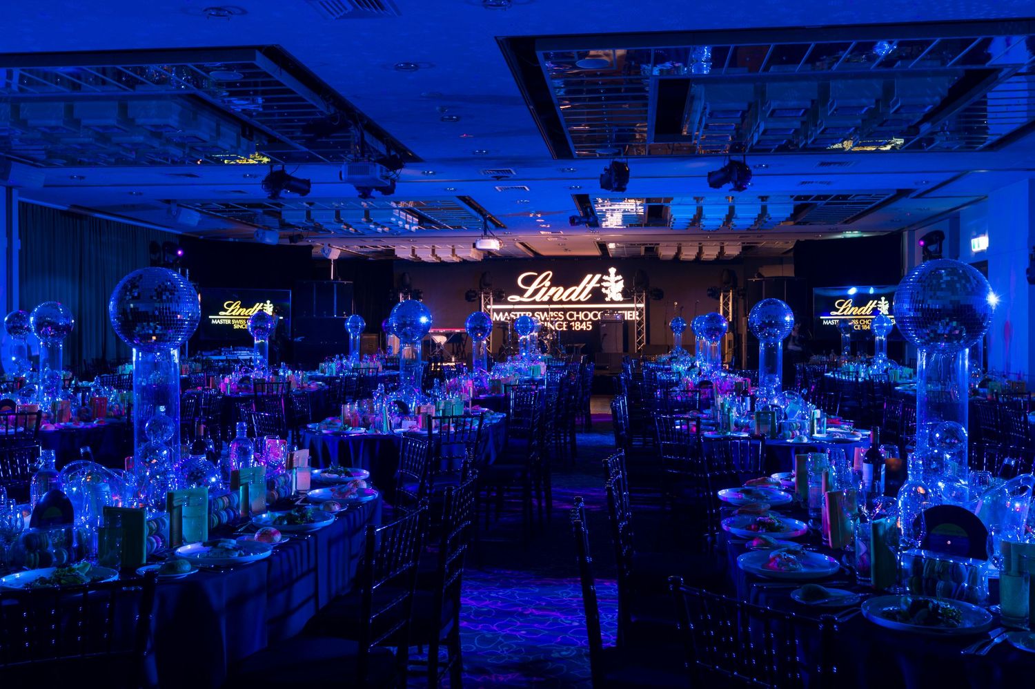 A dimly lit nightclub interior illuminated by blue lighting shows empty tables and chairs in a panoramic view.