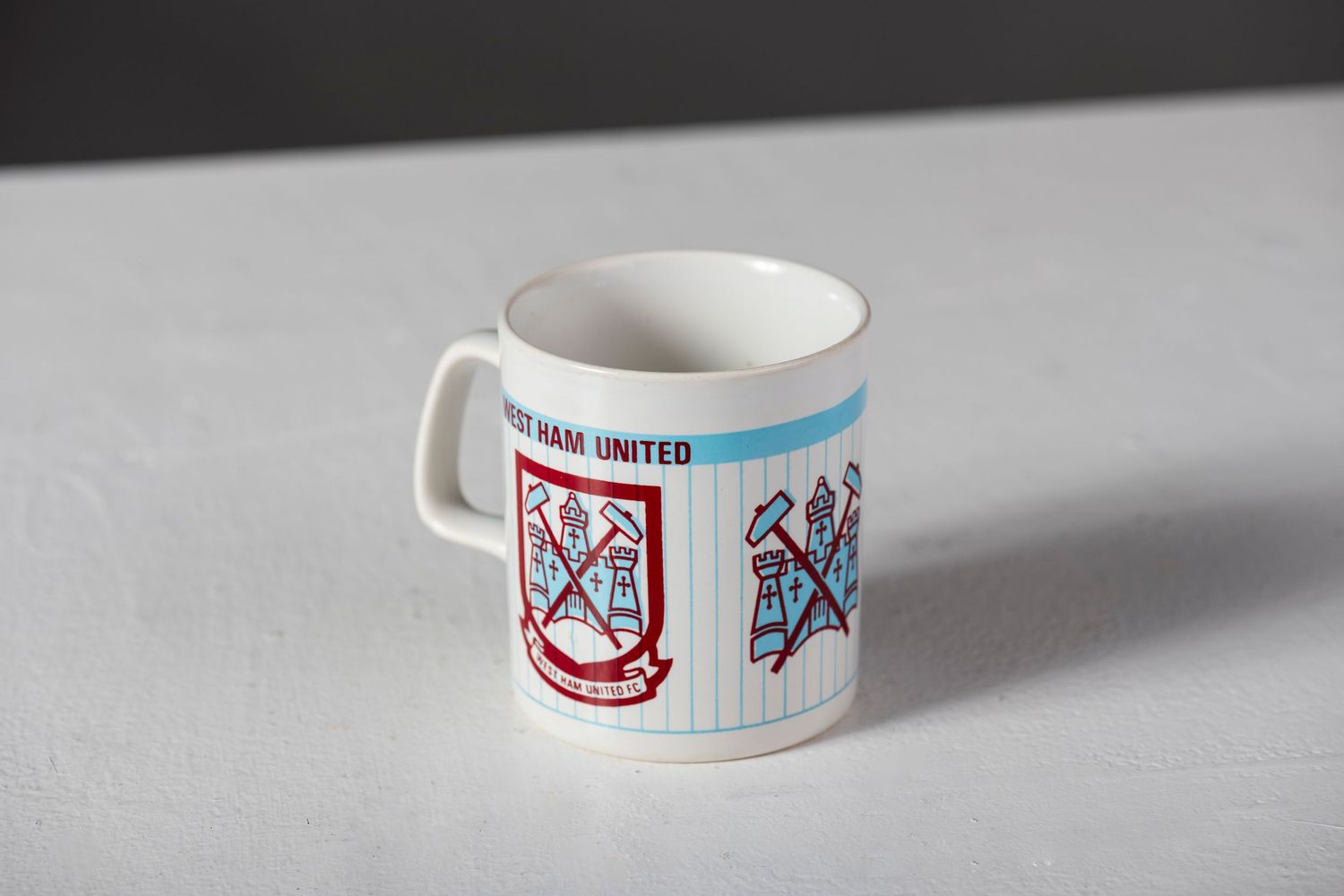 A white ceramic coffee mug featuring vintage sports team logos or crests in red and blue colors on a gray surface.