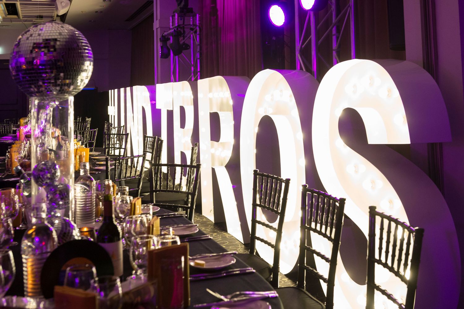 Large illuminated white letters displayed at an elegant sponsored styled event setup with disco ball and chairs in purple lighting.