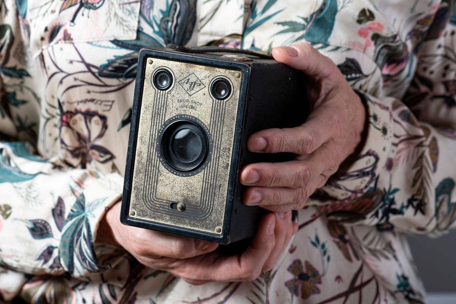 Vintage box camera with brass details held in hands against floral patterned fabric background.