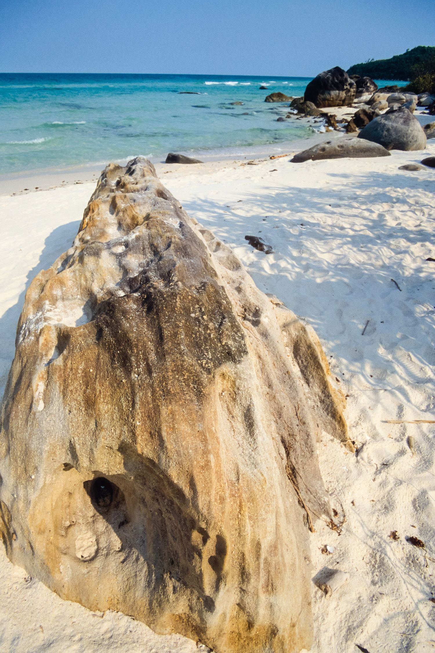 Large rock formation on a white sandy beach with turquoise ocean waters in the background.