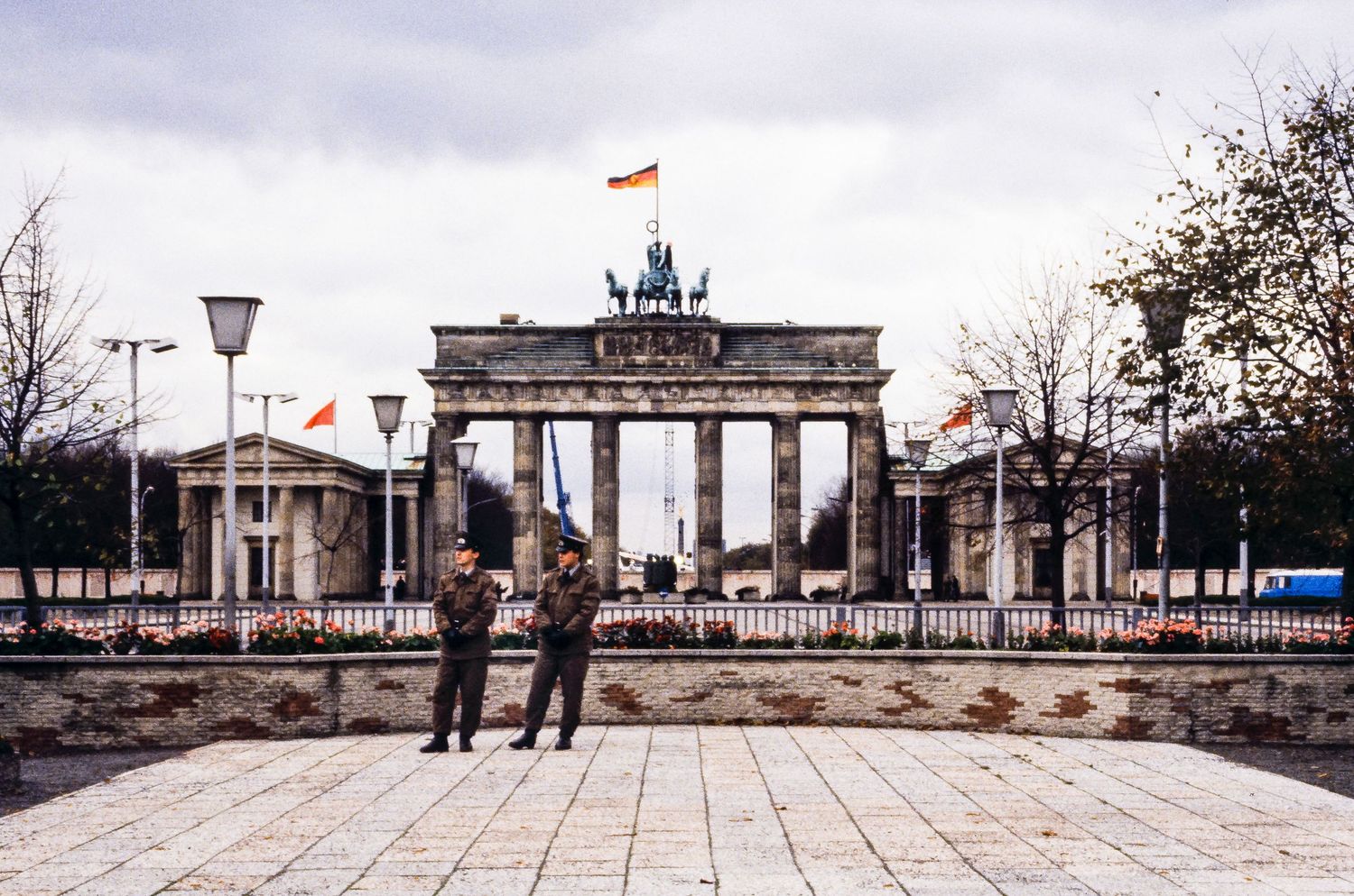 Panoramic view of the Brandenburg Gate in Berlin with German flags flying against a cloudy sky.