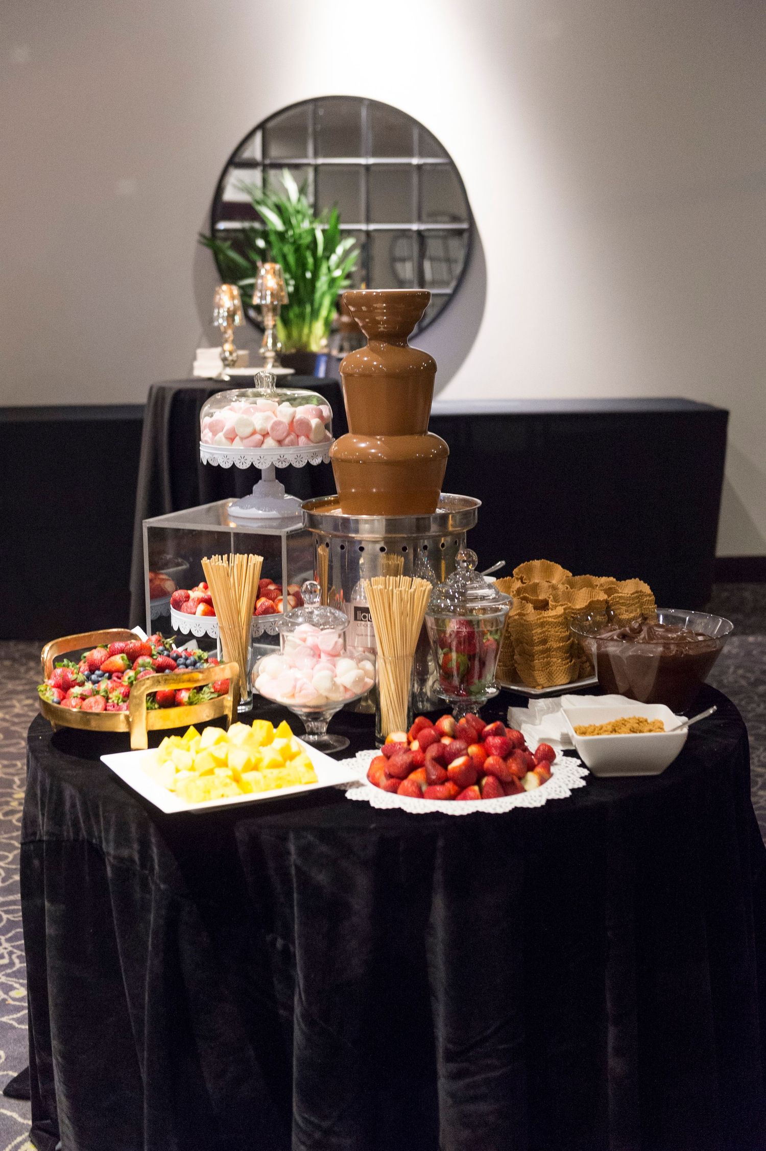 A chocolate fondue dessert station with fresh fruits, marshmallows, and dipping items arranged on a black tablecloth.