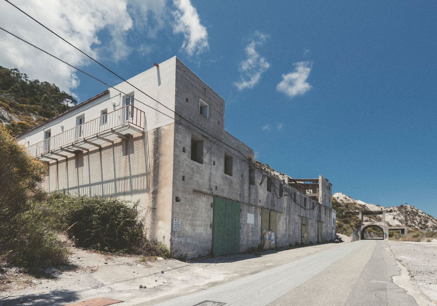 Abandoned concrete industrial building with white walls stands along empty road under blue sky with white clouds.