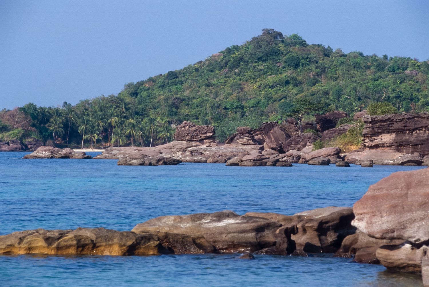 Rocky coastline with turquoise waters and green hills in the background under blue skies.