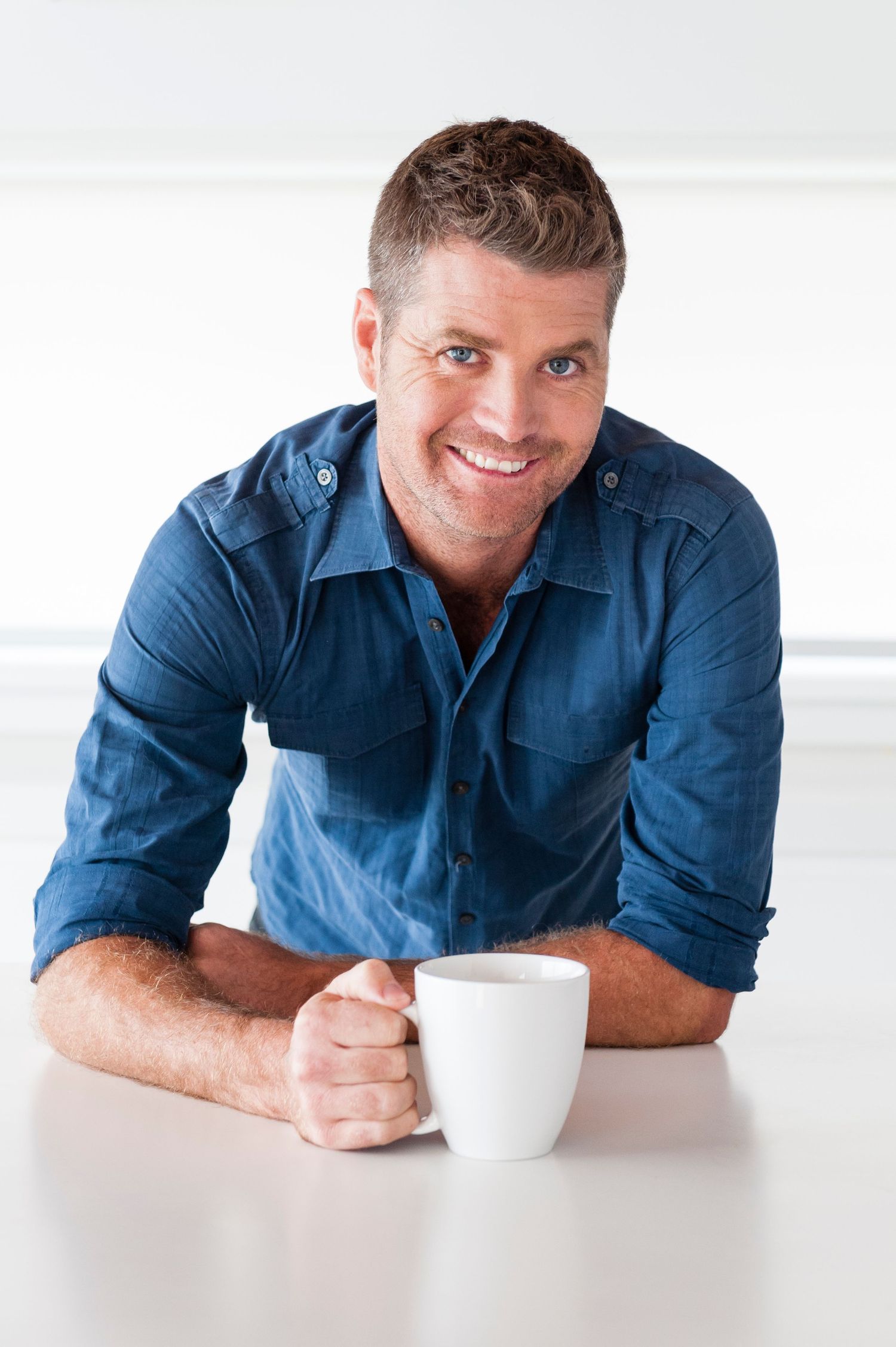 Professional wearing a blue shirt smiles while leaning on a white counter with a coffee cup.