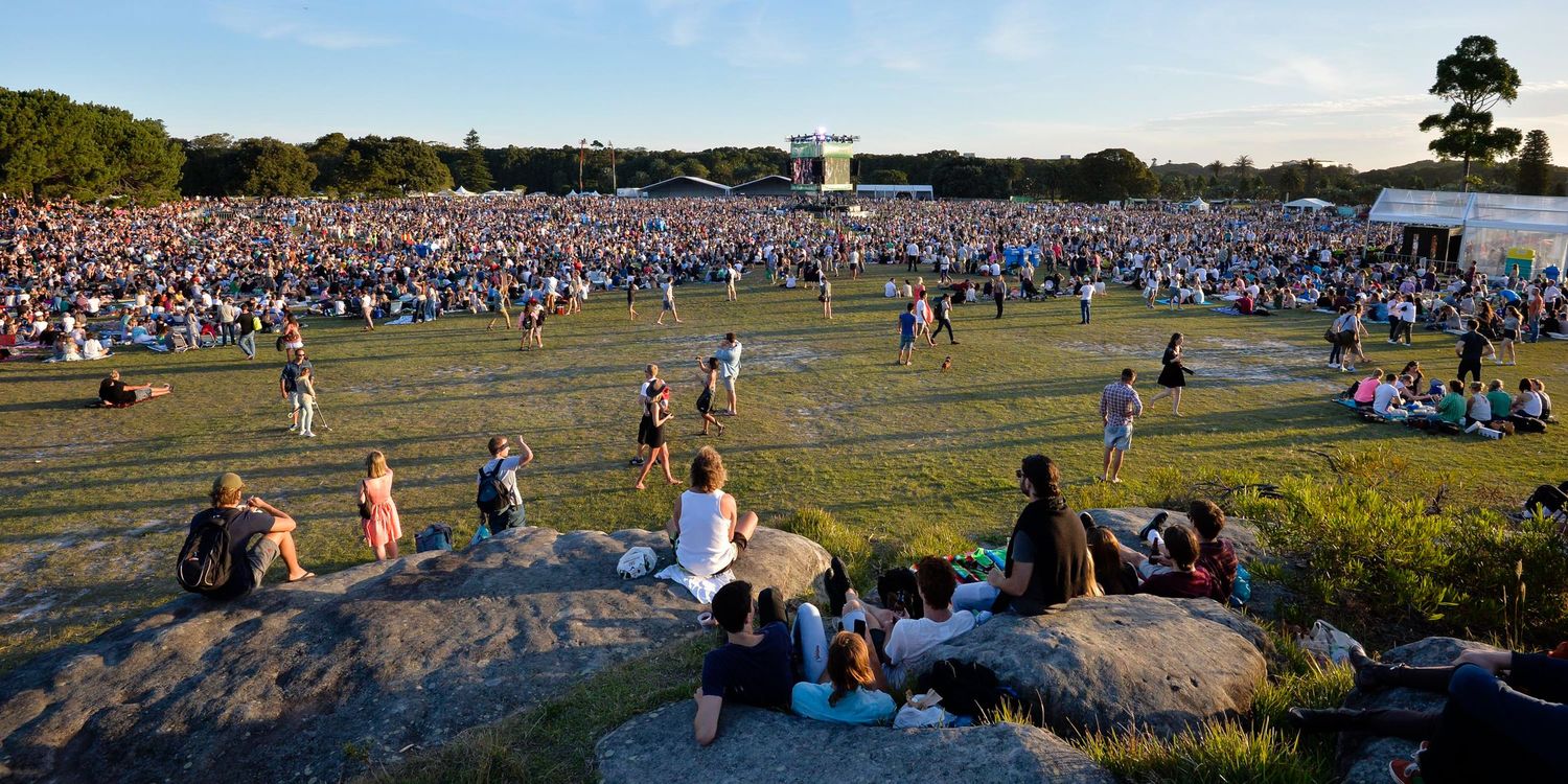 A panoramic view shows crowds of people gathering on a grassy field during an outdoor event on a sunny day.