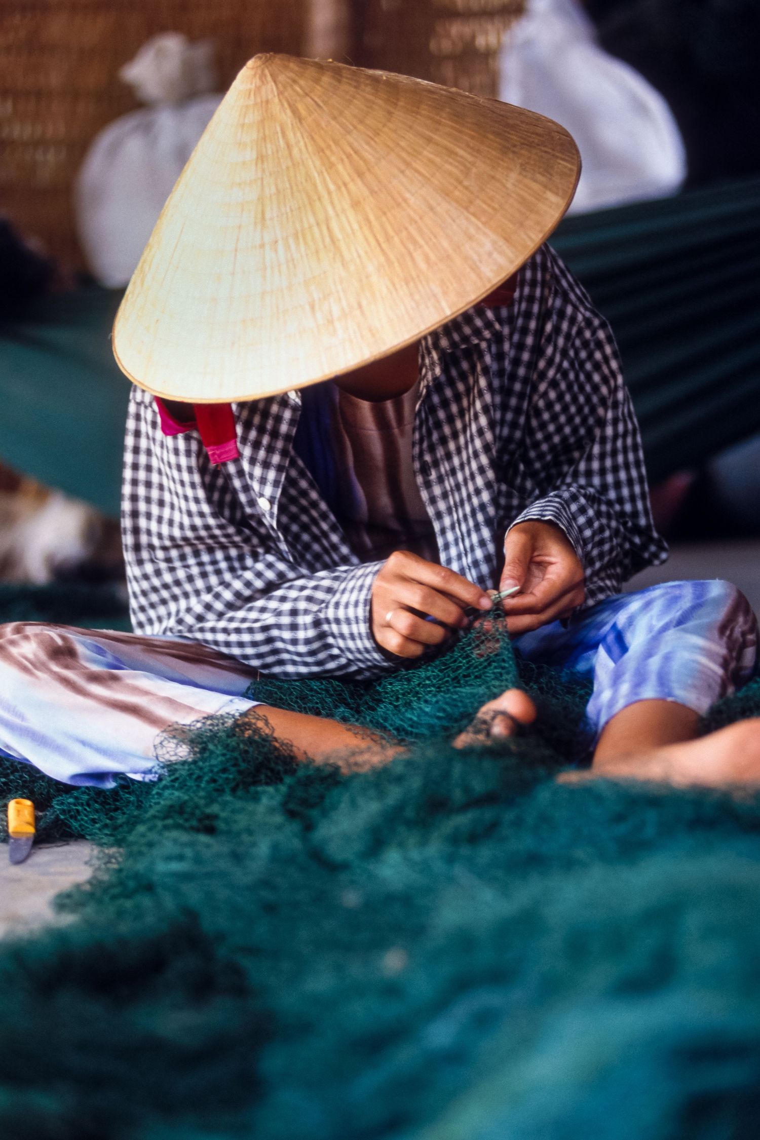 Person wearing traditional conical hat mending fishing nets in Vietnam.