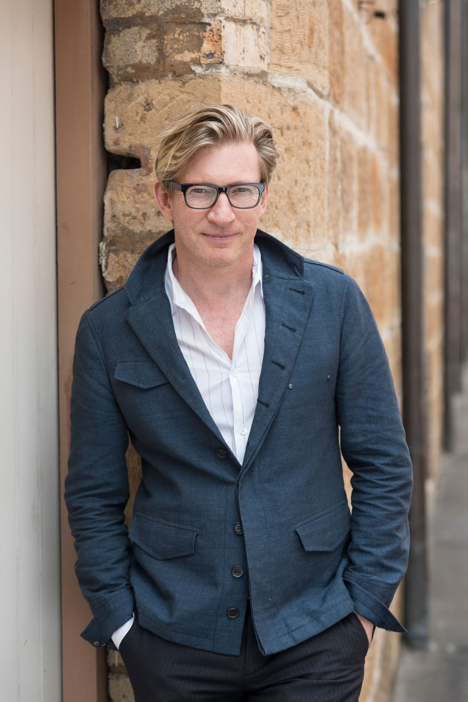 Professional in navy blazer and glasses poses casually against stone wall.