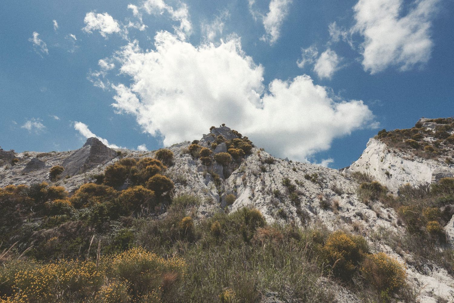 Snow-capped mountain peaks rise against a bright blue sky with scattered white clouds in a panoramic landscape view.