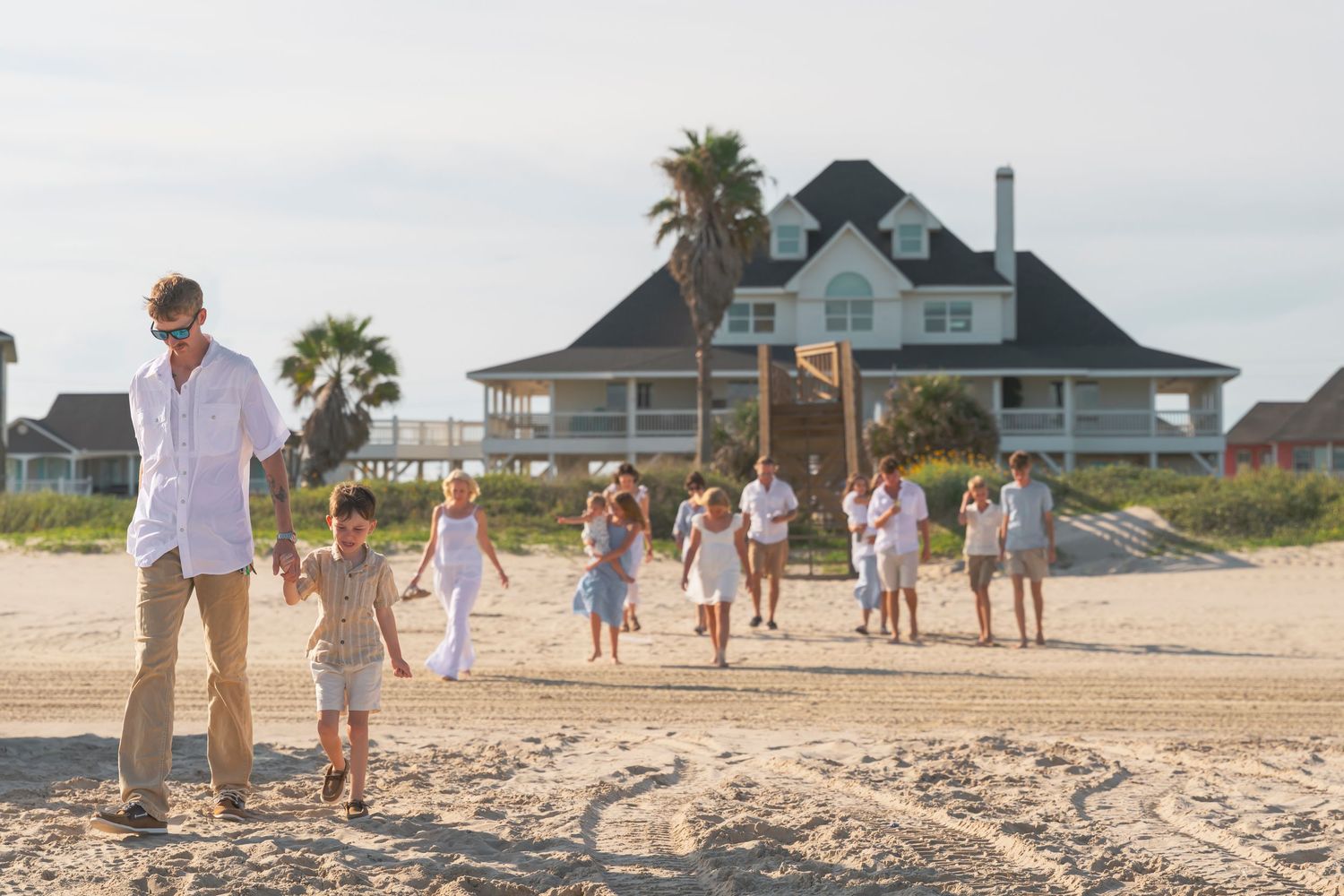 Large family group walking on a sandy beach near a coastal house on a sunny day.