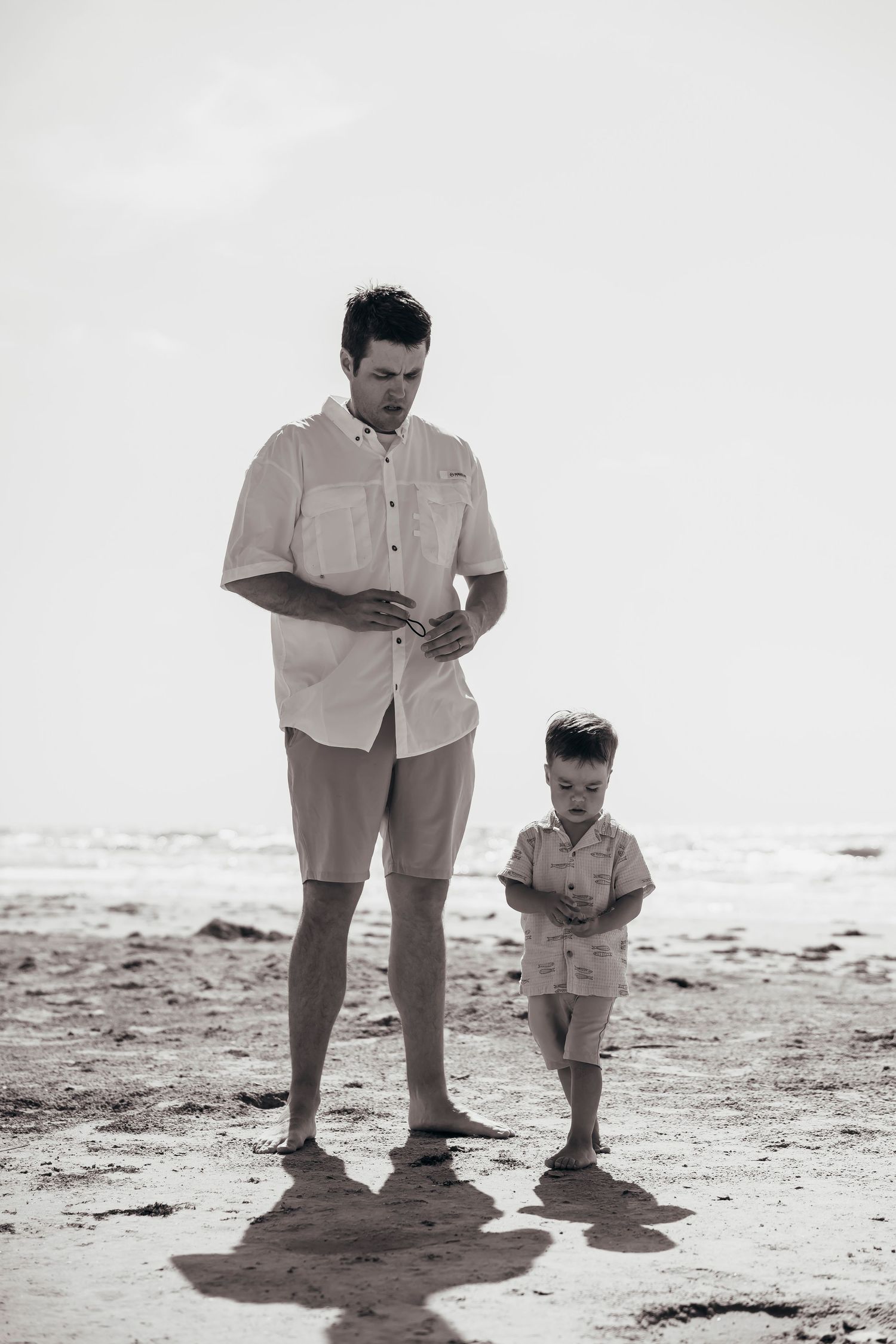 Father and young son standing barefoot on sandy beach in black and white photo, both looking down.