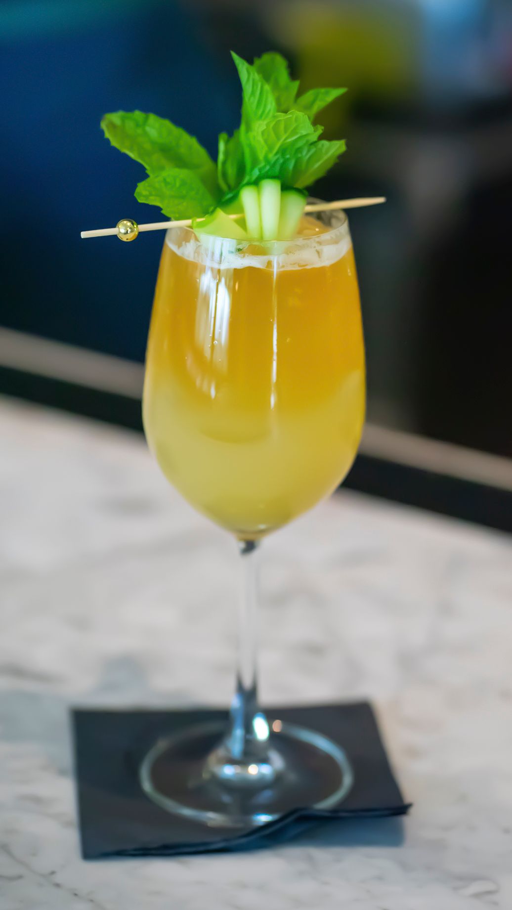 An elegant yellow cocktail garnished with fresh mint and cucumber in a wine glass on a marble surface.