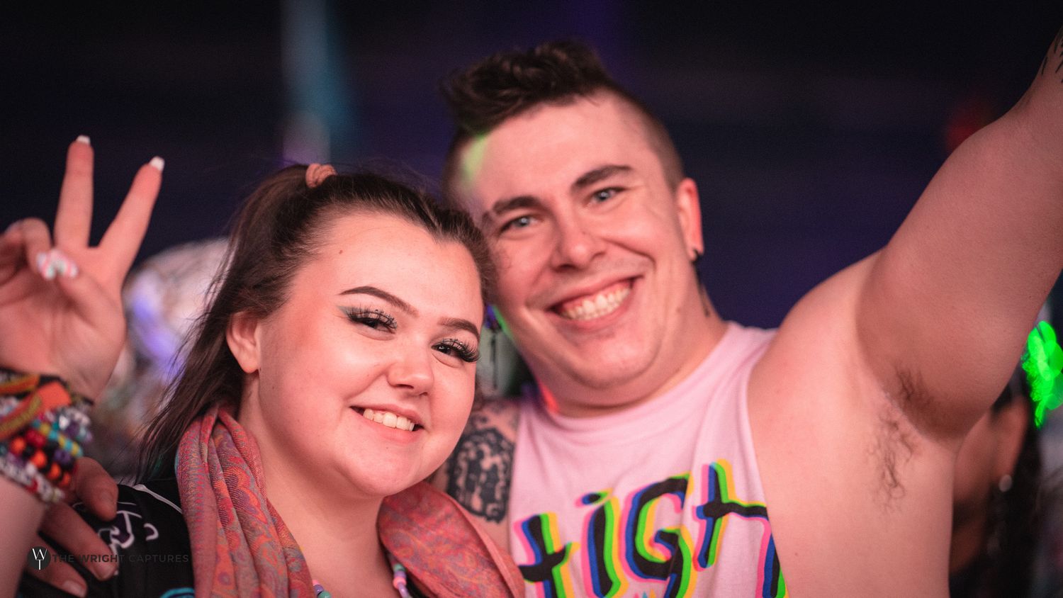 Two smiling friends pose for a selfie at a music festival, one flashing a peace sign.