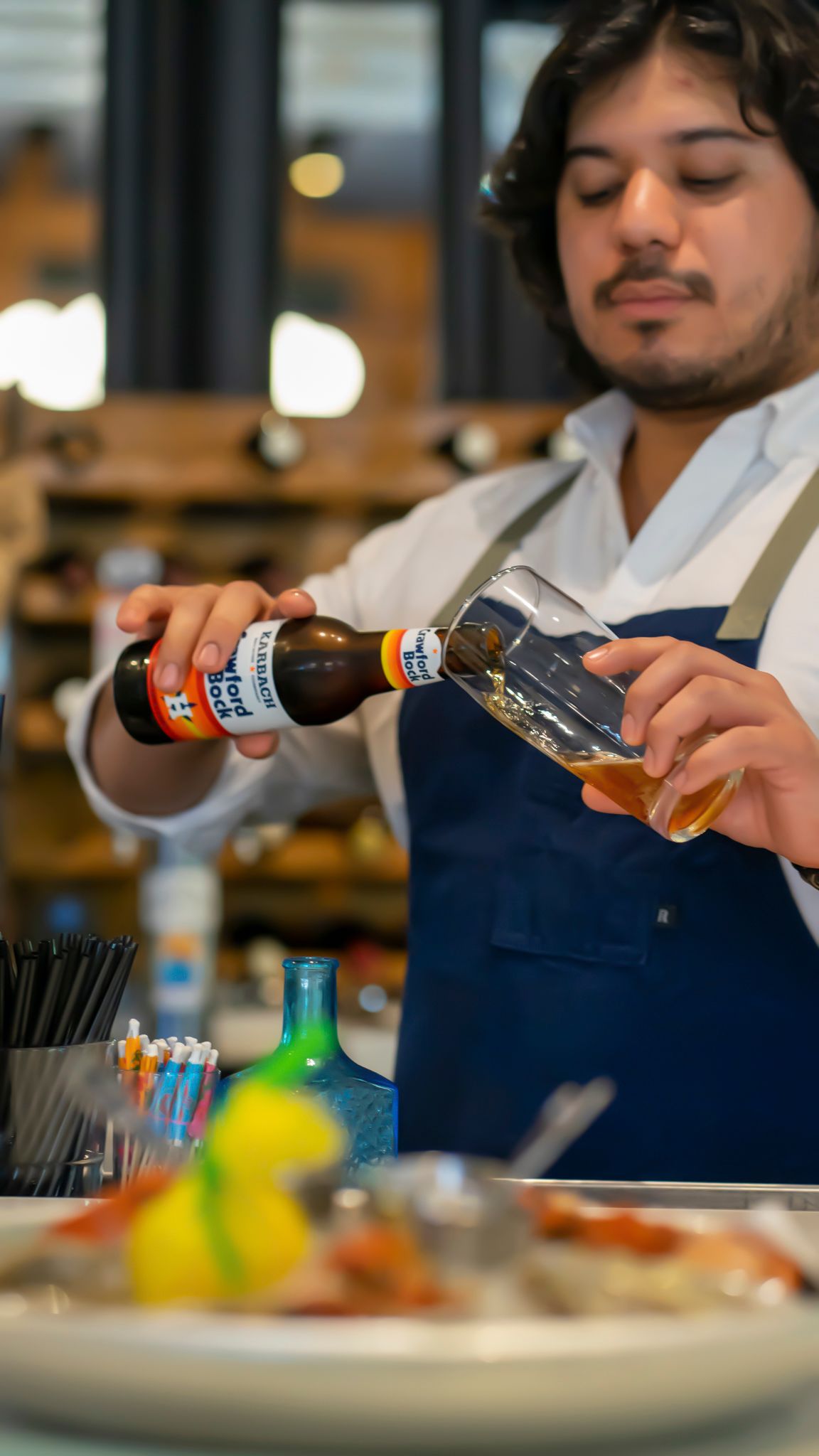 Bartender in blue apron pouring craft beer into tilted glass at restaurant bar counter.