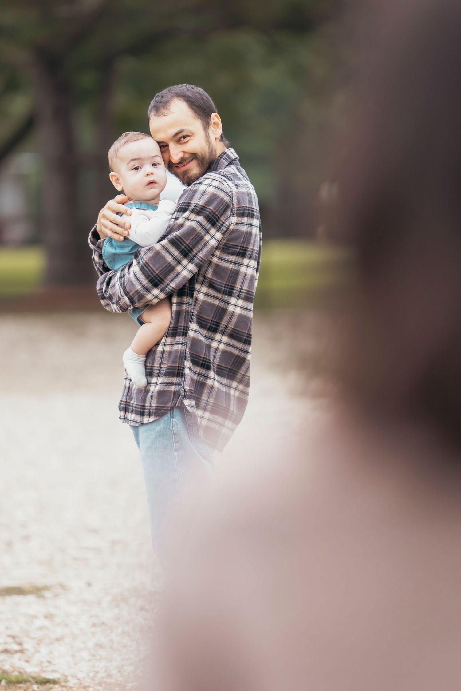 A smiling father holding his baby in a park, wearing a plaid shirt, sharing a tender moment outdoors.