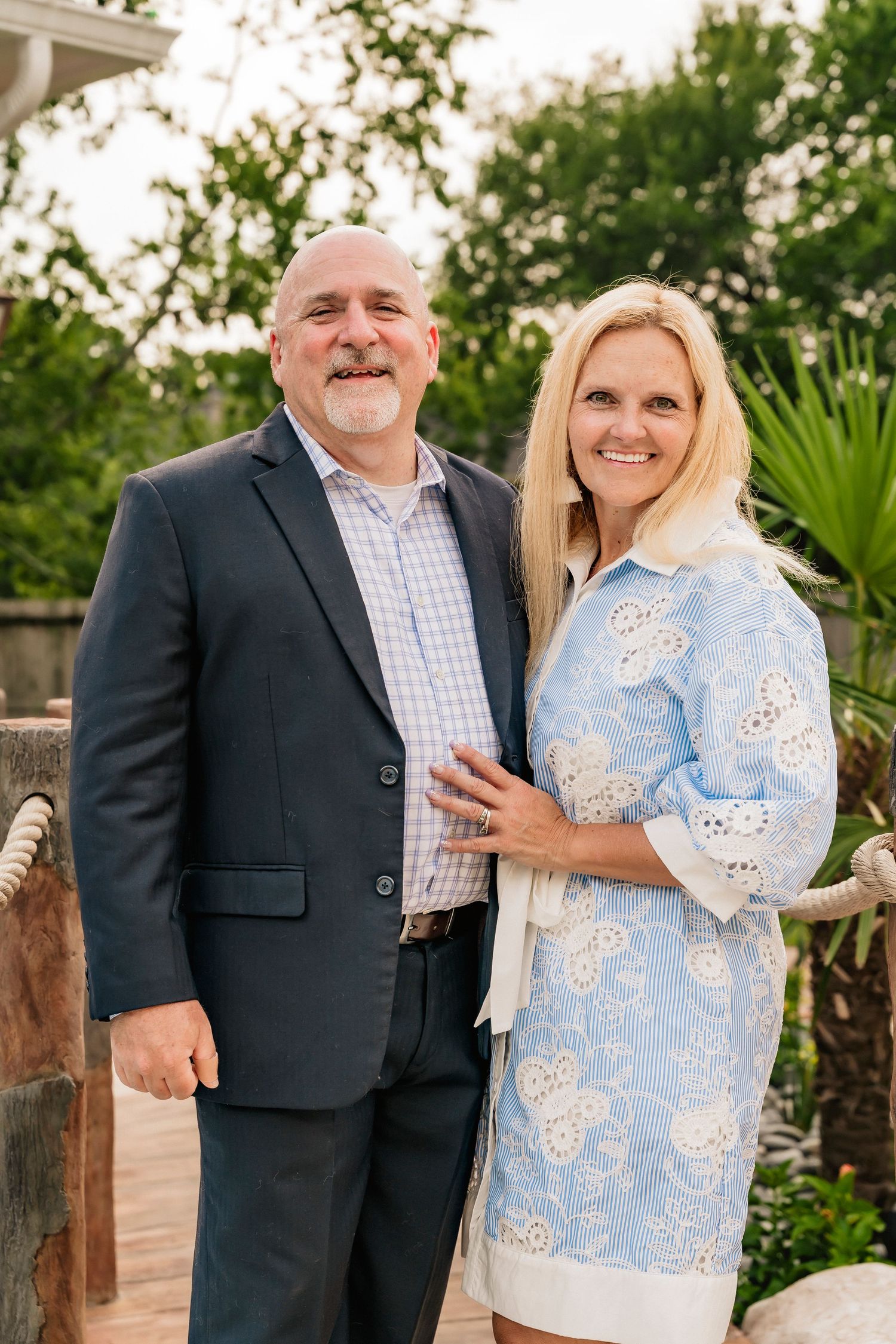 A smiling couple poses outdoors, man in navy blazer and woman in light blue floral dress, with lush greenery behind them.