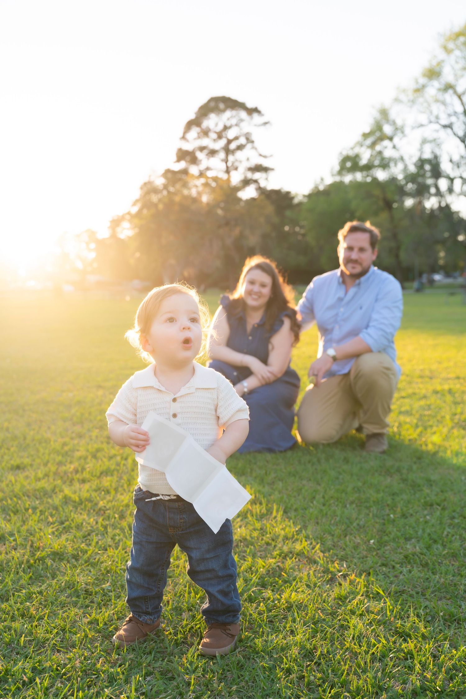 A toddler holds a paper airplane in a sunny park while parents sit on green grass in the golden hour background.